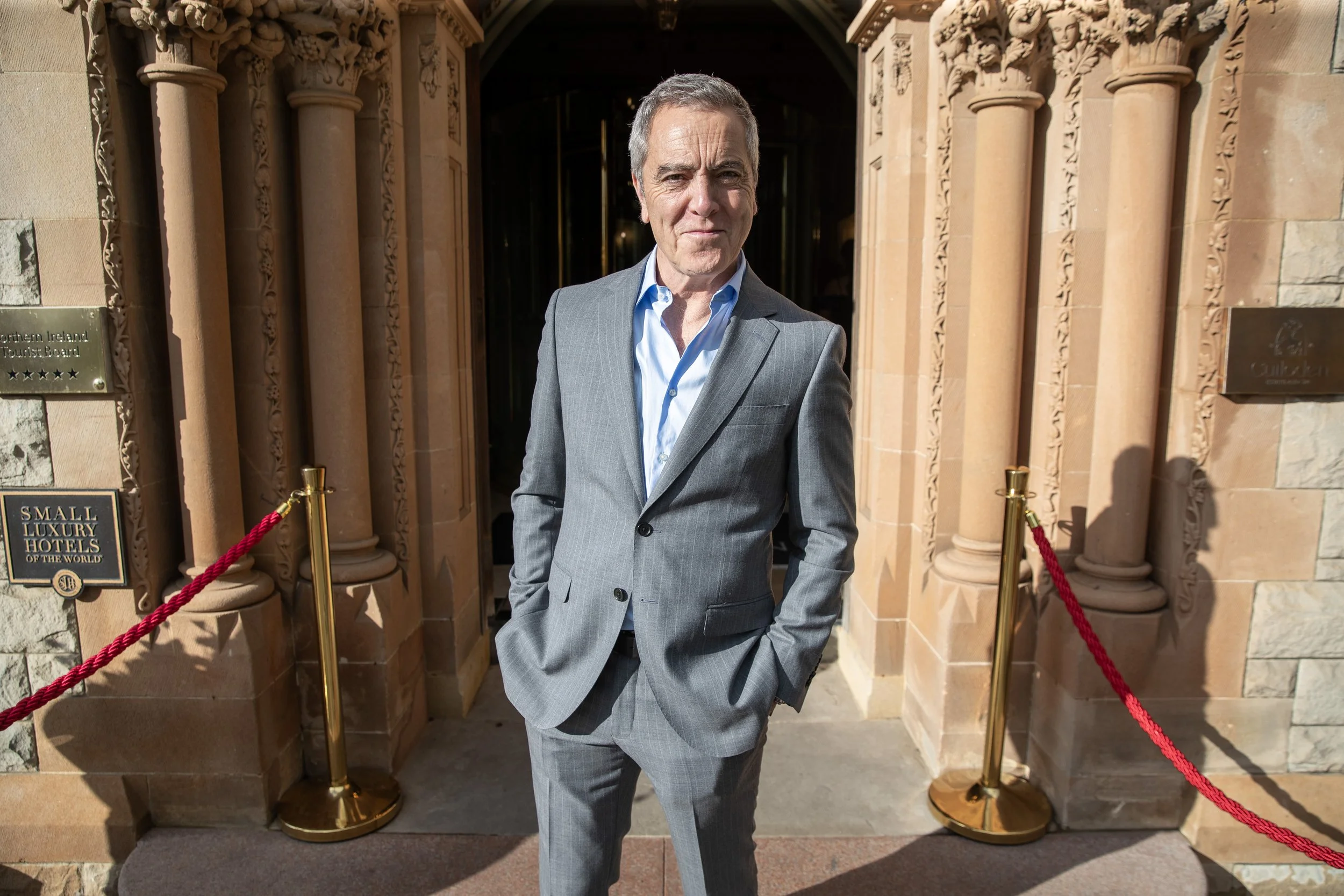 A man in a gray suit standing in front of the entrance to a hotel, with a red rope barrier to the side.