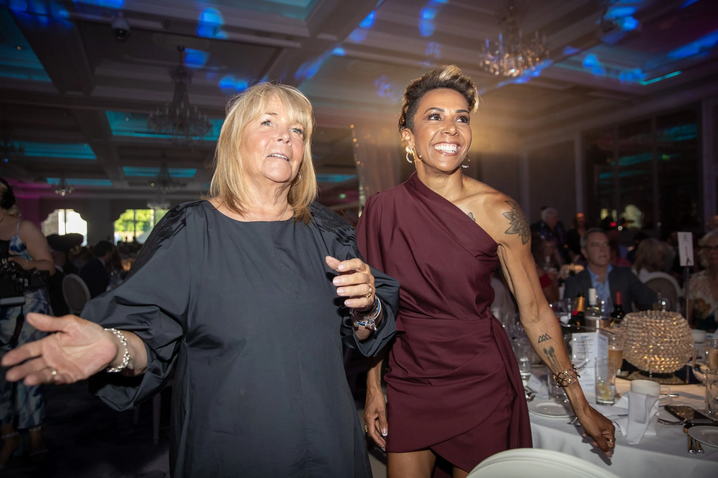 Two women standing together at a formal event, smiling and wearing elegant dresses. The background shows a decorated banquet hall with tables and other guests.