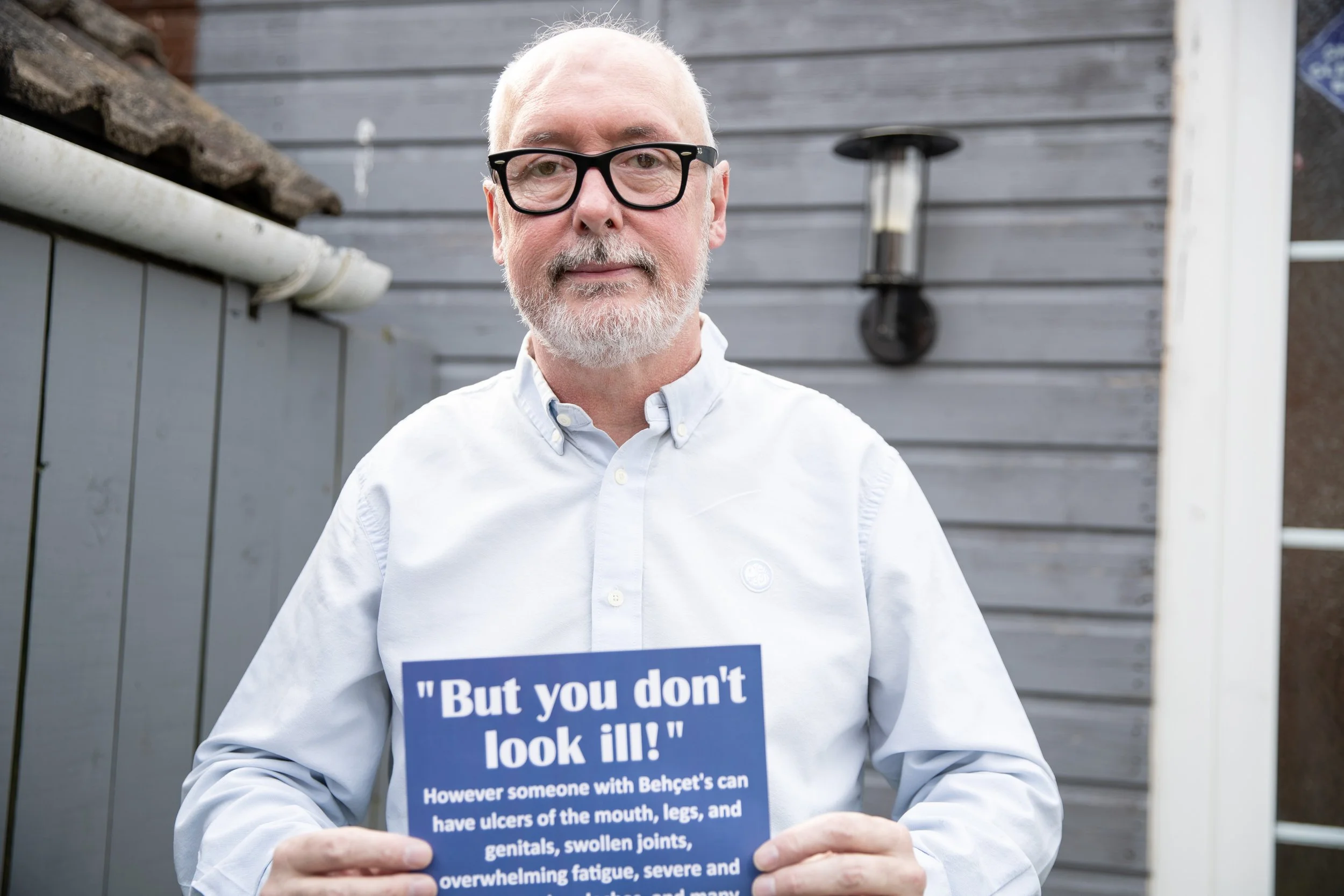 An older man with white hair, beard, glasses, wearing a white button-up shirt, holding a blue sign with white text about Behçet's disease, standing outdoors in front of a gray wooden fence and a wall-mounted outdoor lamp.