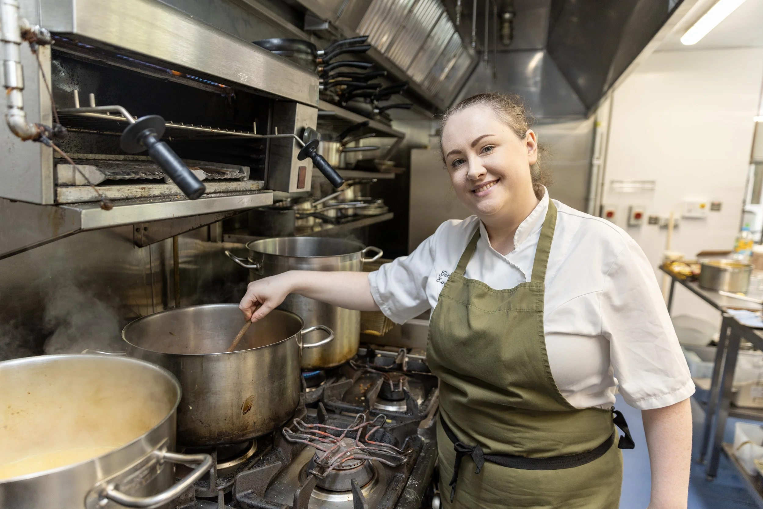A female chef in a white uniform and green apron stirring a pot on a commercial stove in a busy kitchen.