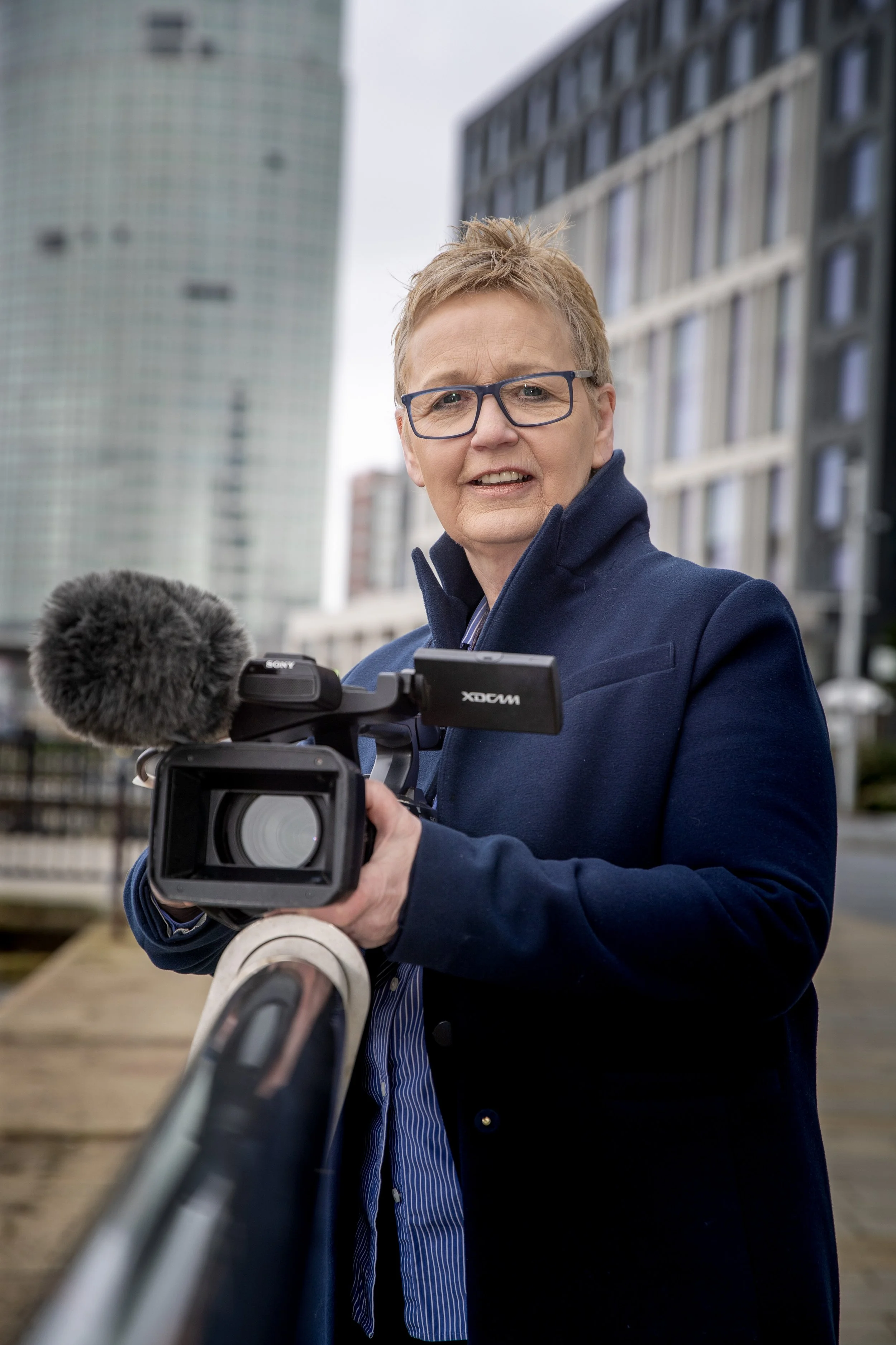 A woman with short hair and glasses holding a video camera outdoors on a city street with modern high-rise buildings in the background.