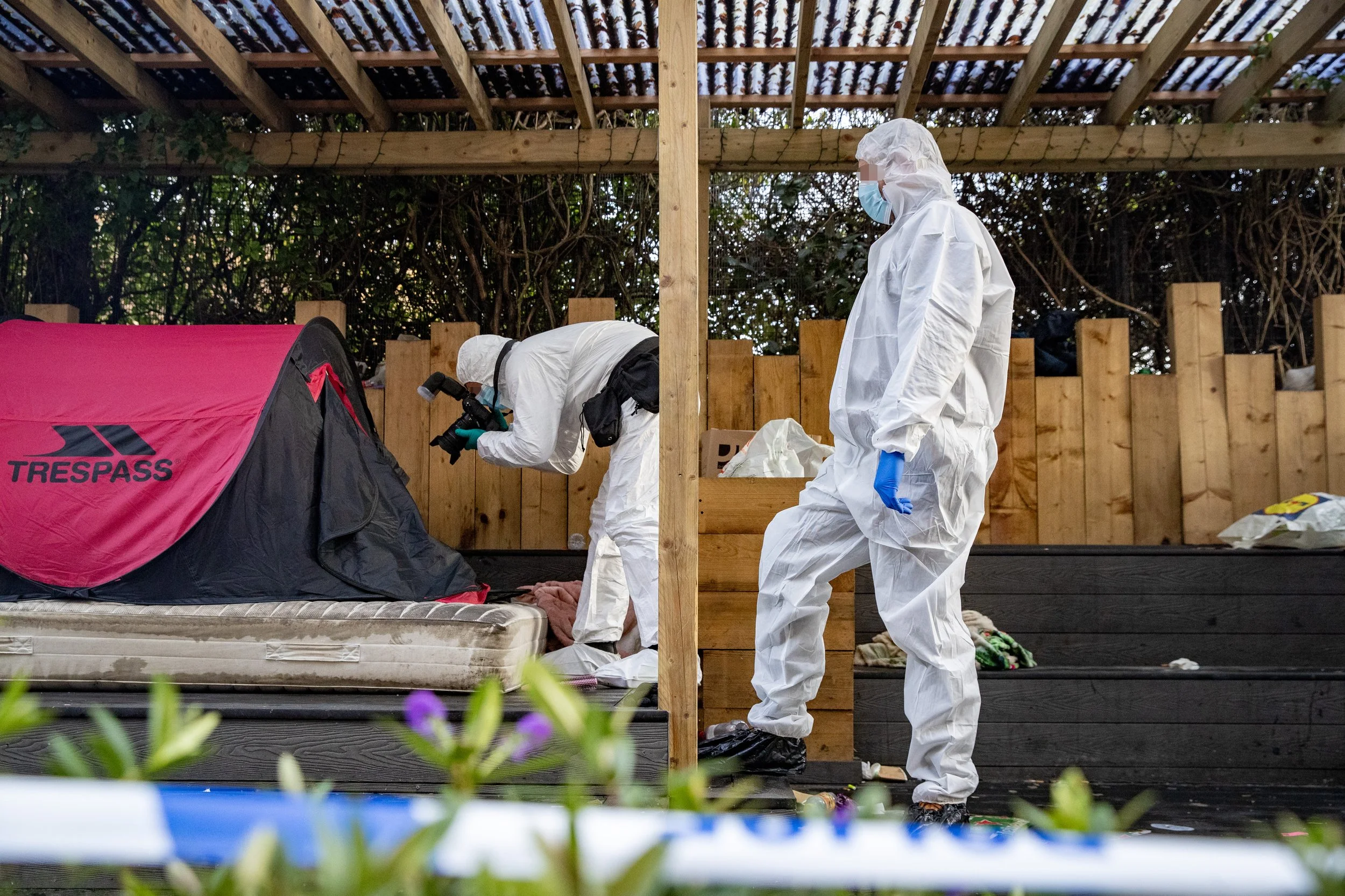 Two forensic investigators in white protective suits and masks examine a pink tent on a mattress outdoors, surrounded by a wooden fence and foliage.