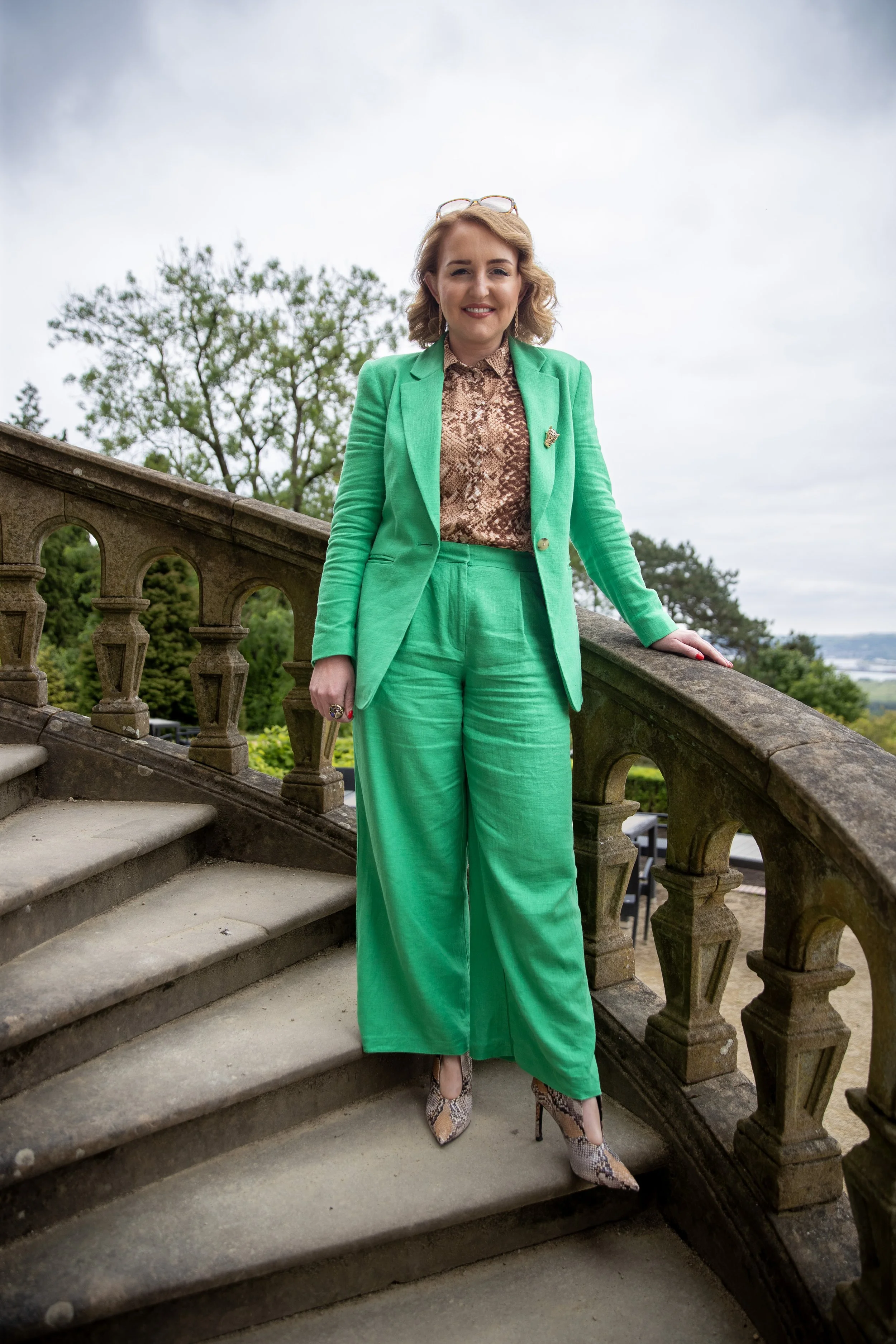 A woman stands on a stone staircase outdoors, wearing a bright green suit, snake-print shoes, and a snake-print blouse. She has short, curly blonde hair and is smiling at the camera.