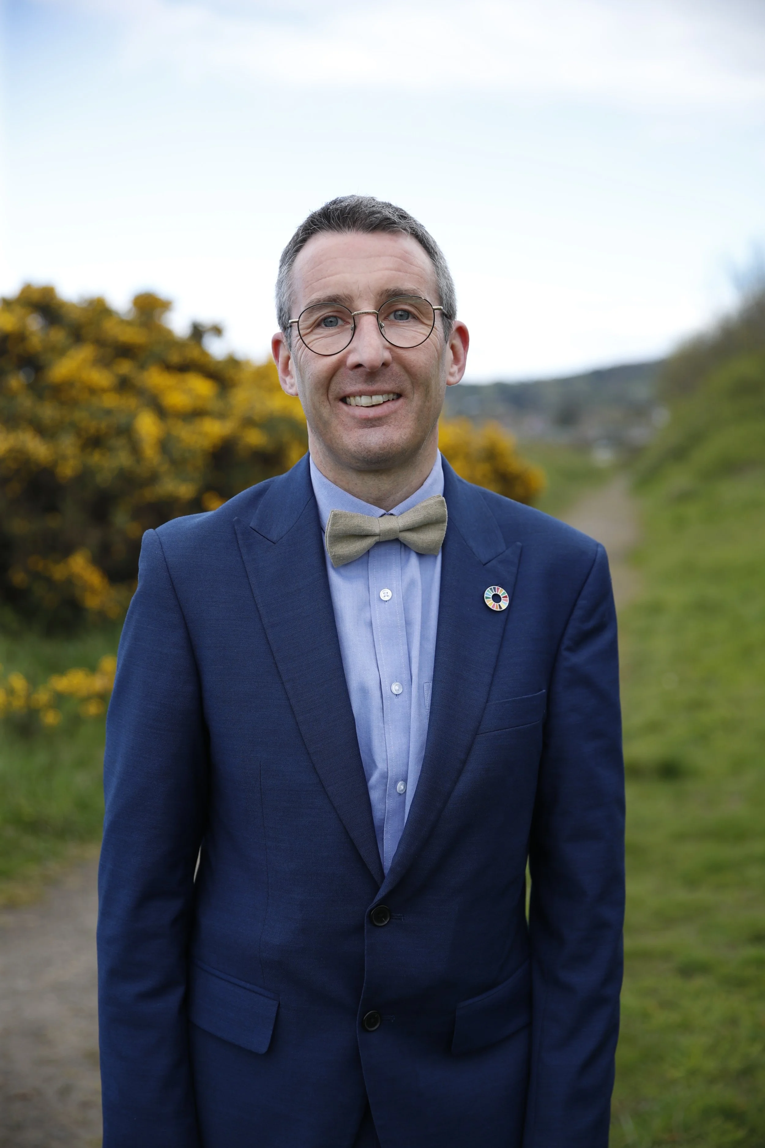 A man in a navy blue suit, light blue shirt, and beige bow tie smiling outdoors, with yellow flowers and greenery in the background.