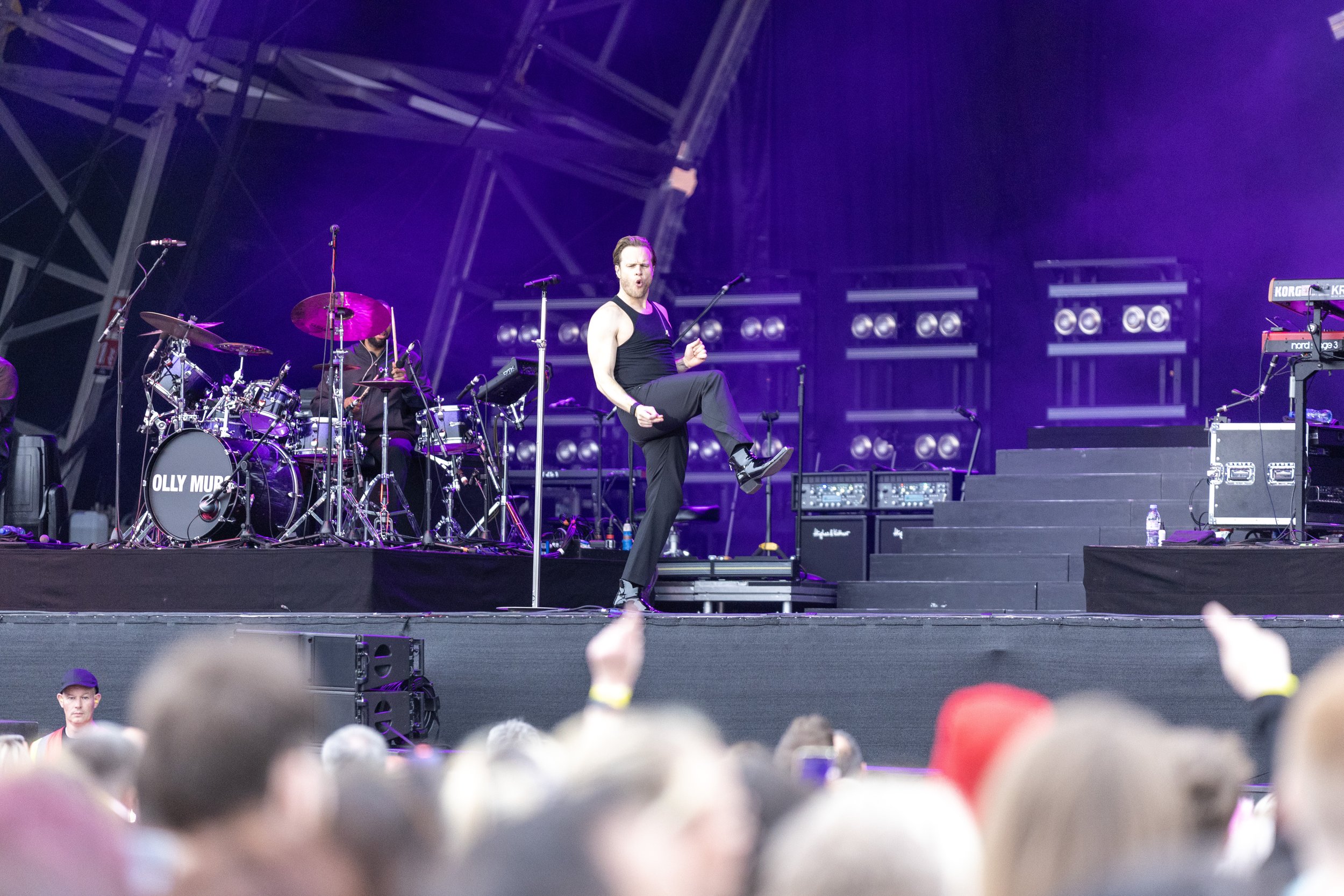 A live concert performance on stage with a male singer in black pants and a tank top, with a drummer and other musical equipment behind him, and an audience in front.