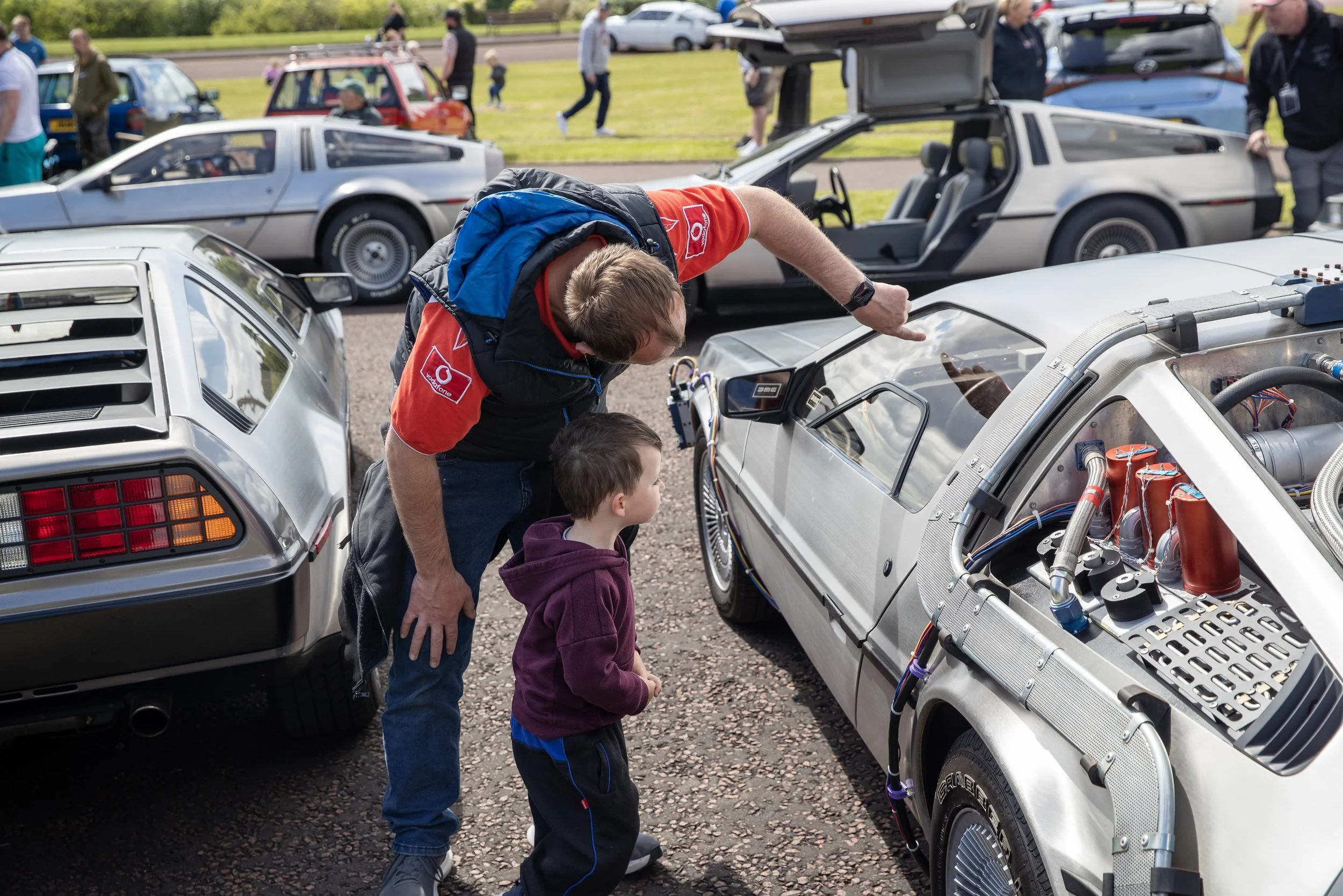 A man and a young boy look at a DeLorean time machine car on display at a car show, with other classic cars and visitors in the background.
