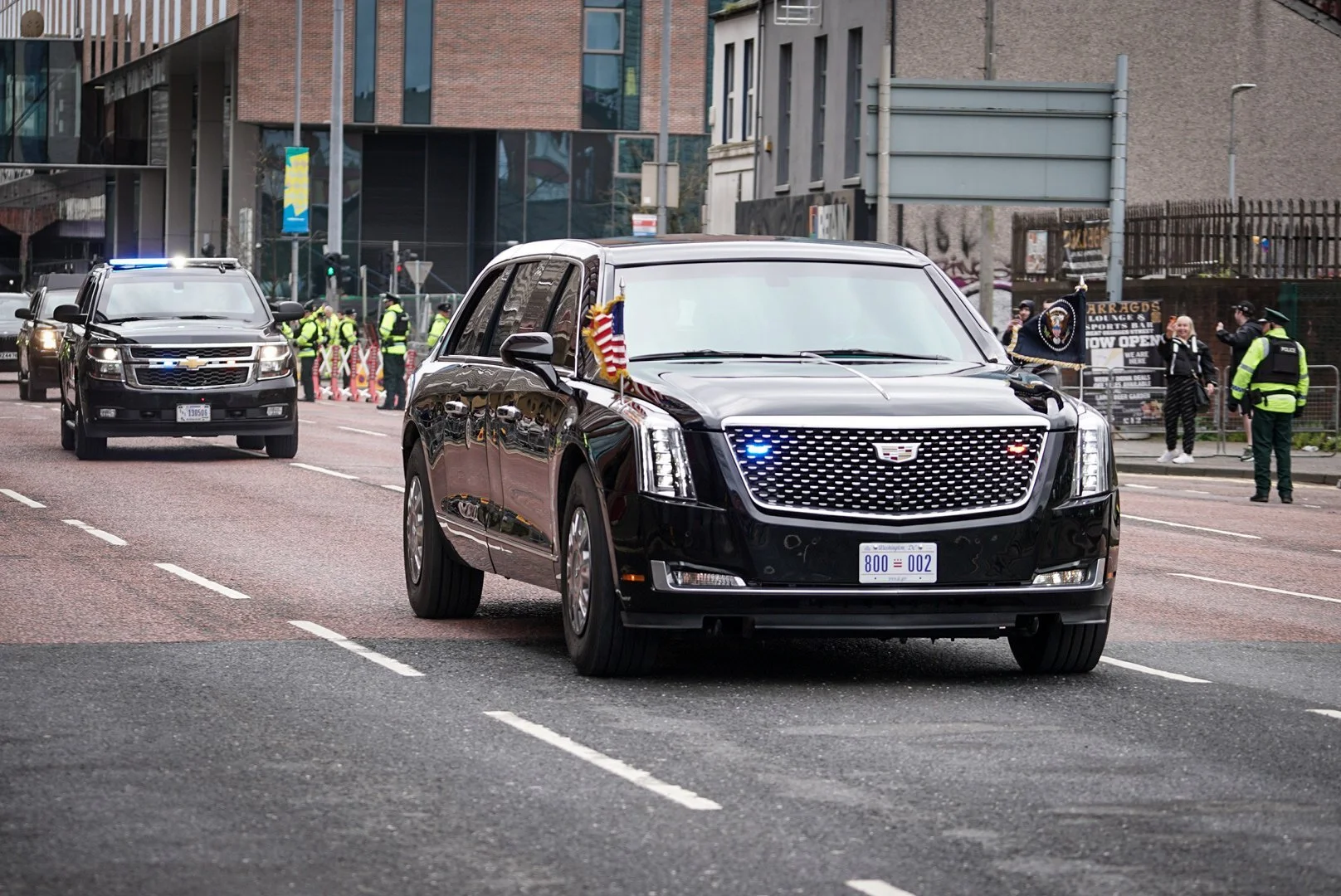 A black Cadillac vehicle leading a procession of police cars on a city street, with officers, onlookers, and barricades in the background.
