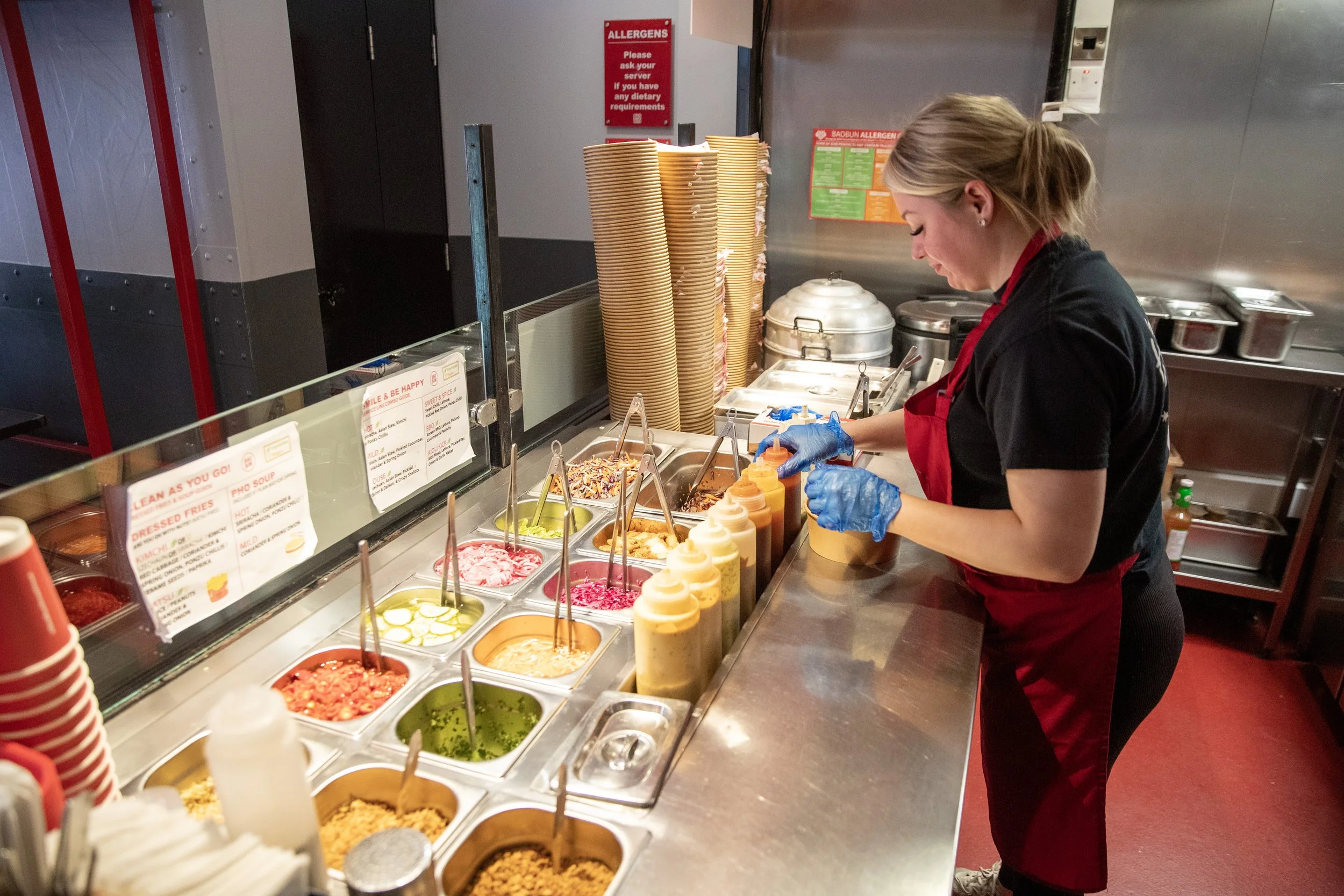 Woman preparing food at a fast-food restaurant counter with various condiments and toppings displayed.