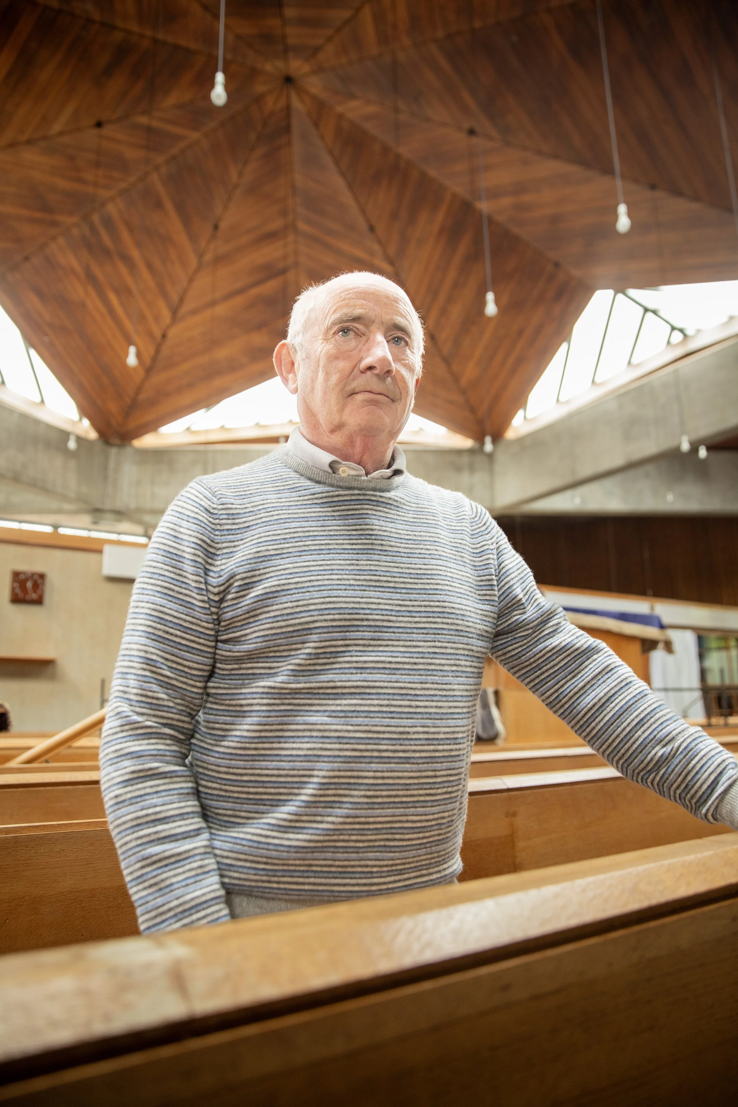 An elderly man in a striped sweater standing in a church pew inside a church with a wooden ceiling and skylights.