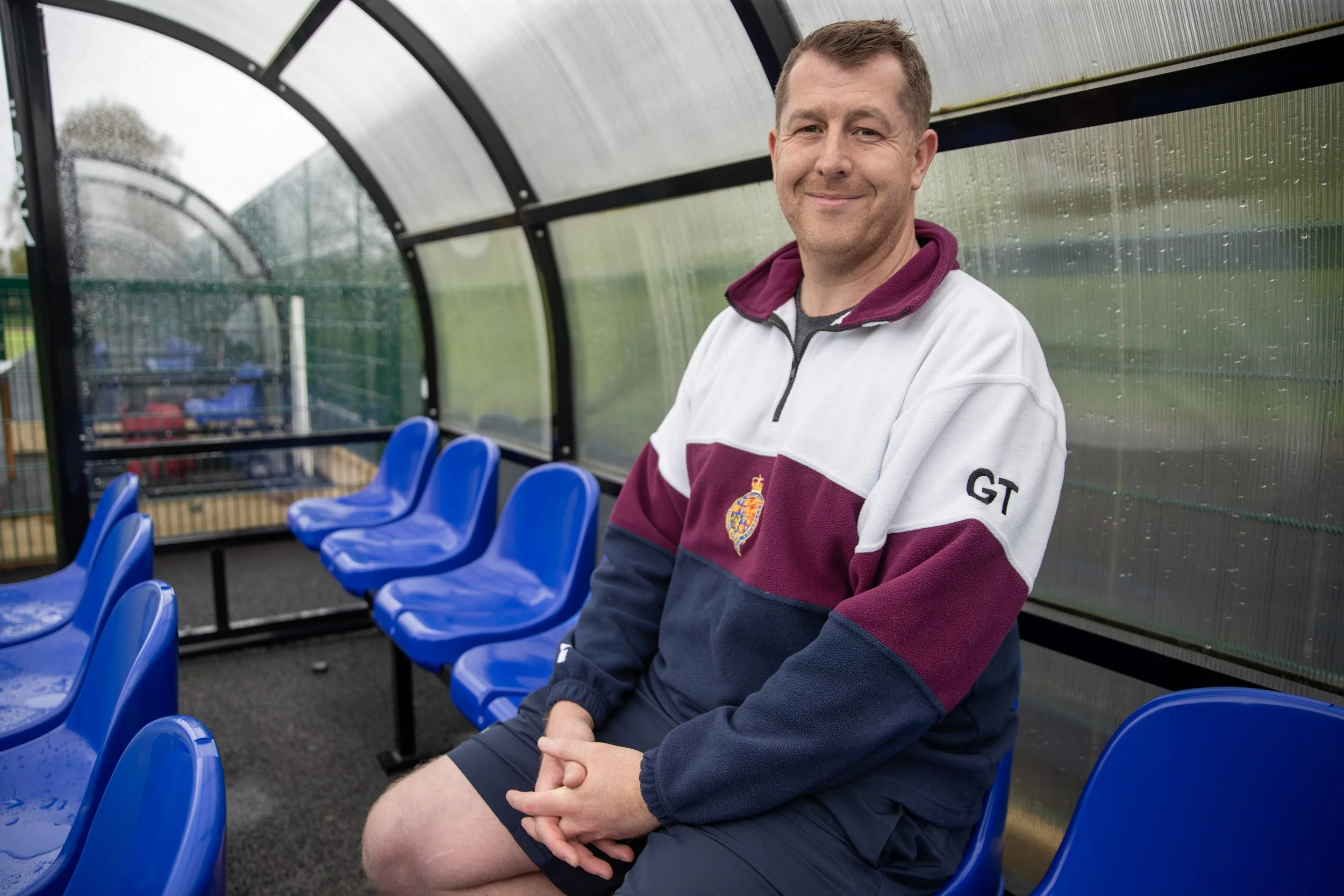 A man sitting on a blue stadium seat inside a covered sports bench area, smiling and wearing a rugby jacket and shorts.
