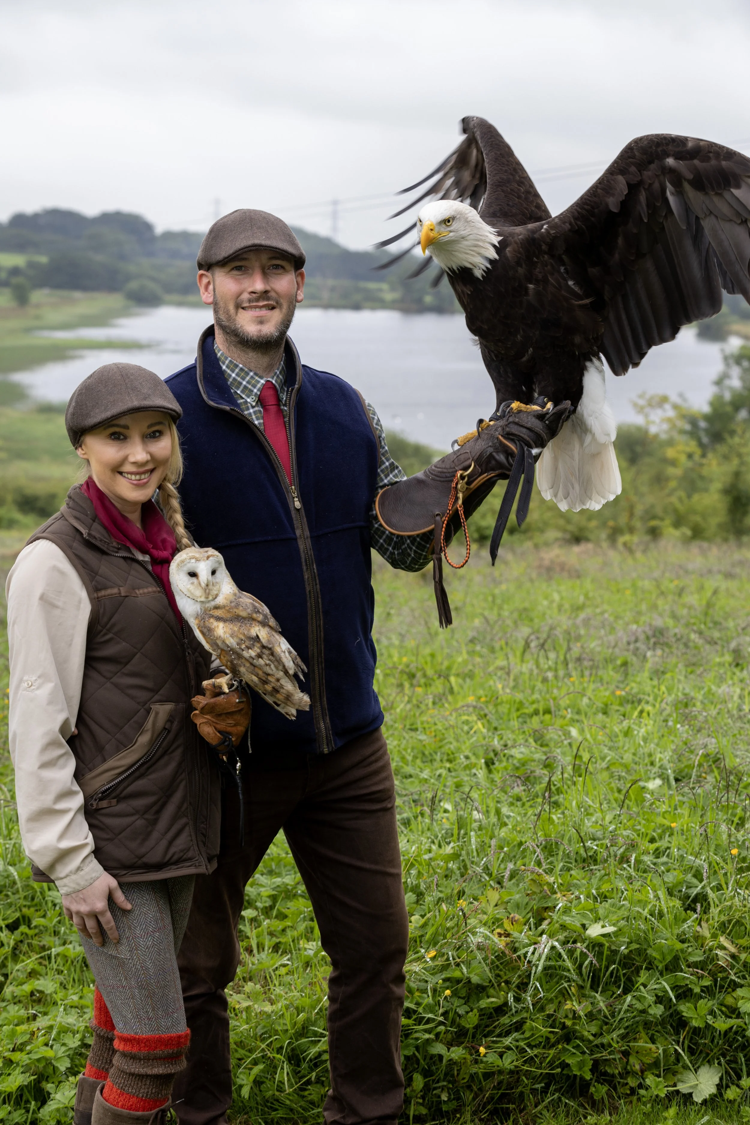 A man and woman dressed in outdoor clothing stand in a grassy field holding two birds of prey, a bald eagle on the man's gloved hand and a barn owl on the woman's gloved arm, with a lake and hilly landscape in the background.