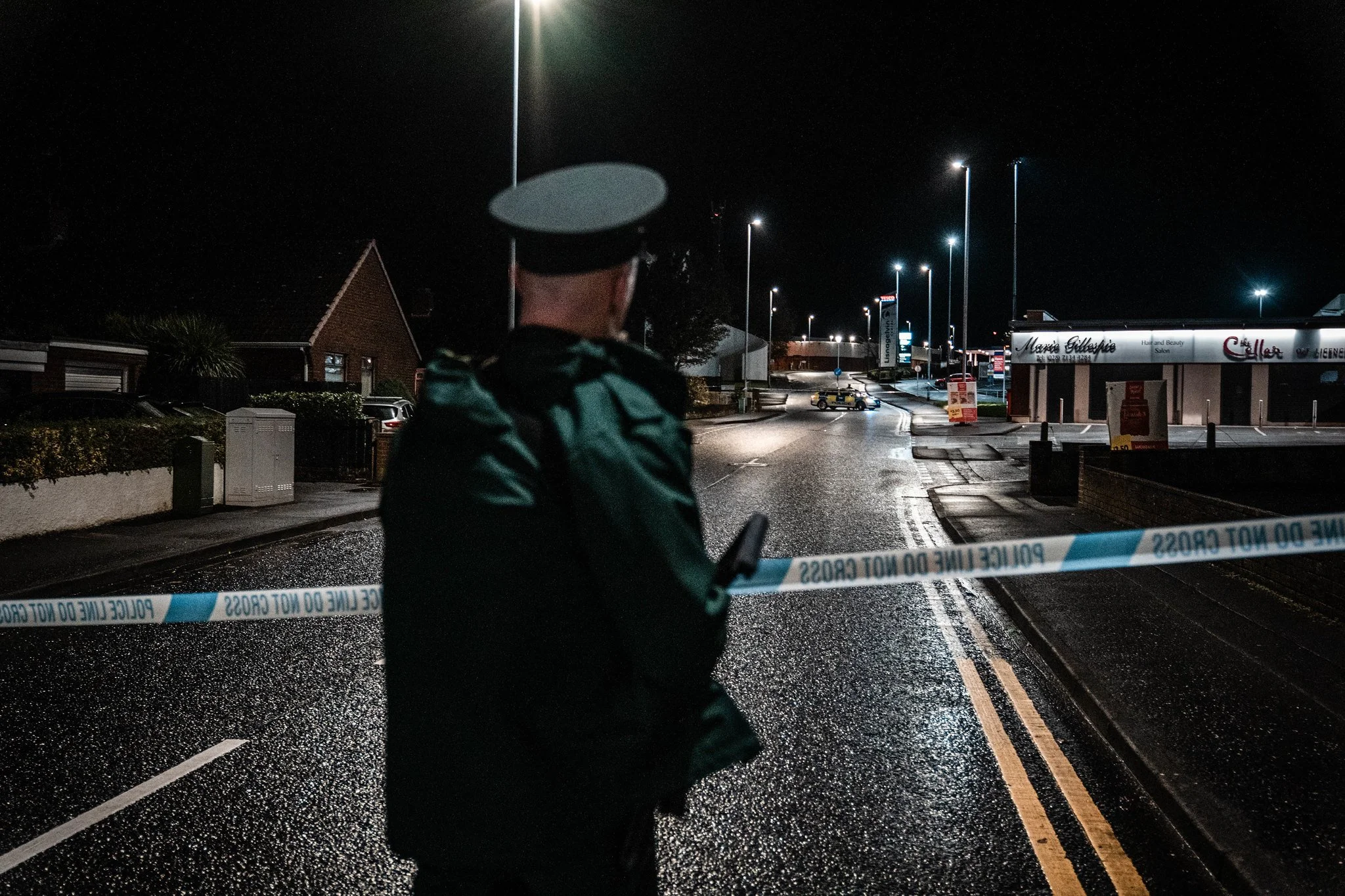 A police officer stands behind police tape on a wet street at night, with streetlights illuminating the area and a small building visible in the background.
