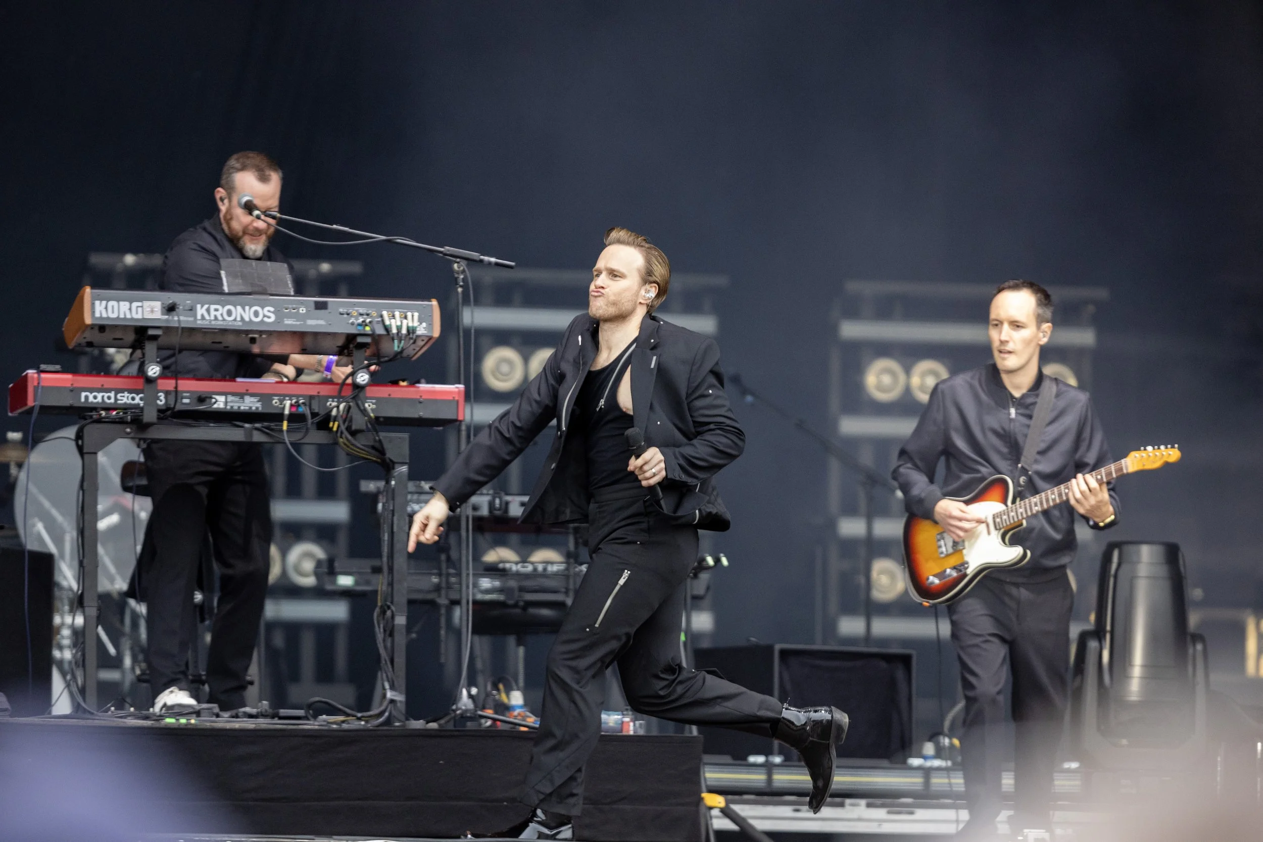 Three male musicians performing on stage, one playing keyboard, one singing with a microphone, and the third playing electric guitar, dressed in dark clothing.