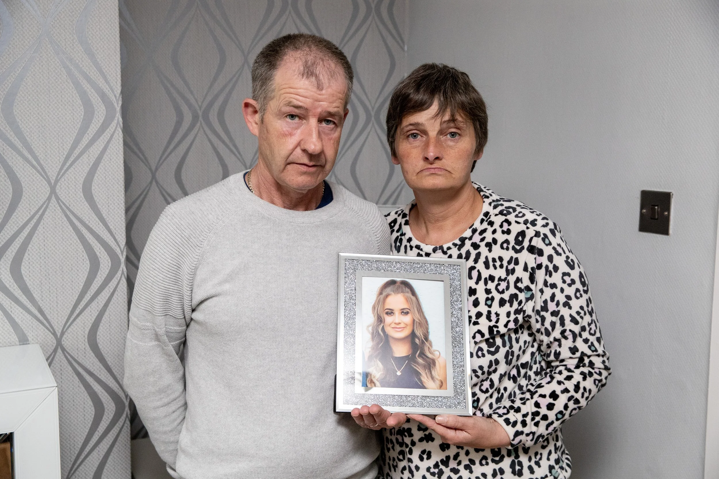 A man and woman stand together indoors, holding a framed photo of a young woman with long, wavy hair, both looking serious.