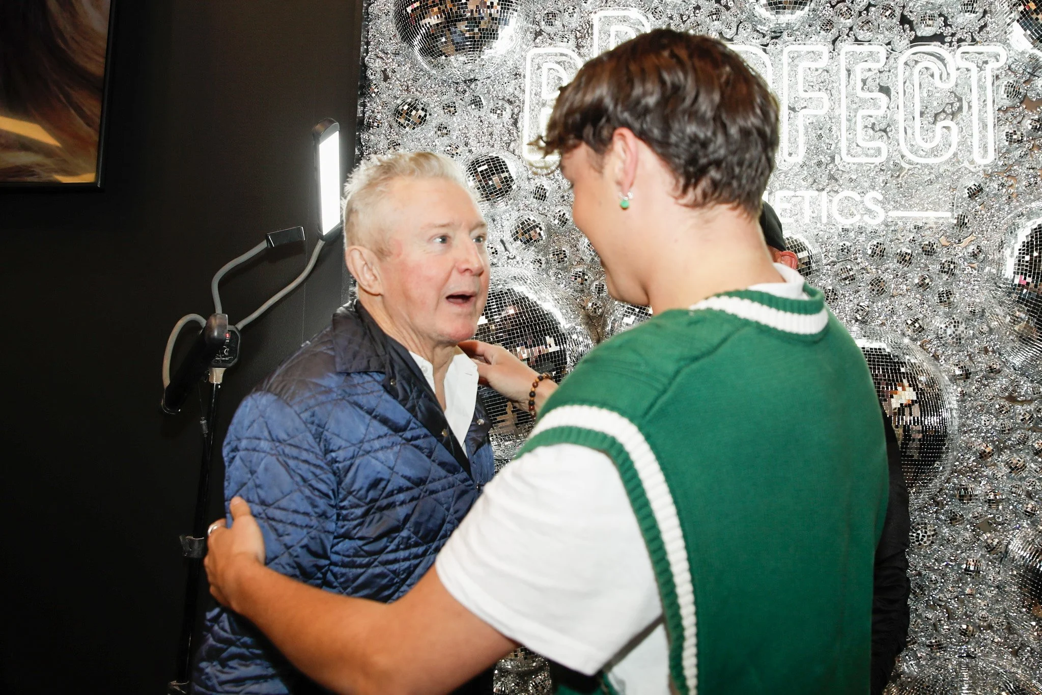 Two people engaged in conversation in front of a shiny, disco ball-themed background with the words 'PERFECT' visible. One person is an older man with light-colored hair, wearing a quilted blue jacket, and the other is a younger person with short dar