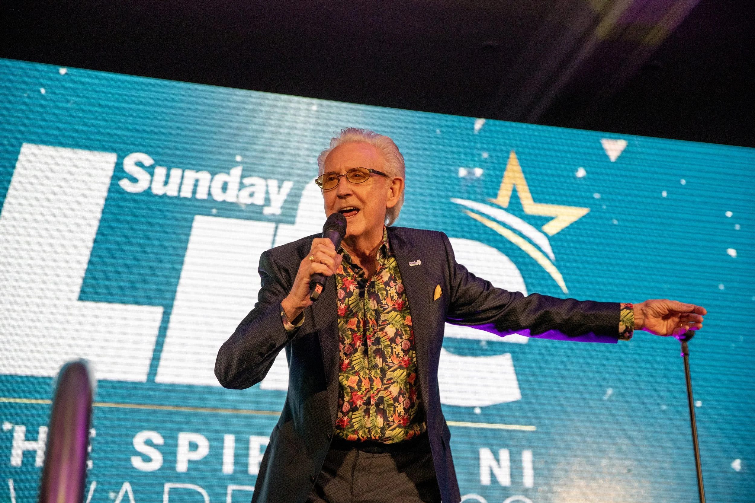 An elderly man with white hair and glasses, wearing a dark blazer and a colorful floral shirt, speaking into a microphone on stage. Behind him is a large screen displaying the word 'Sunday' and a star logo, suggesting a conference or event setting.