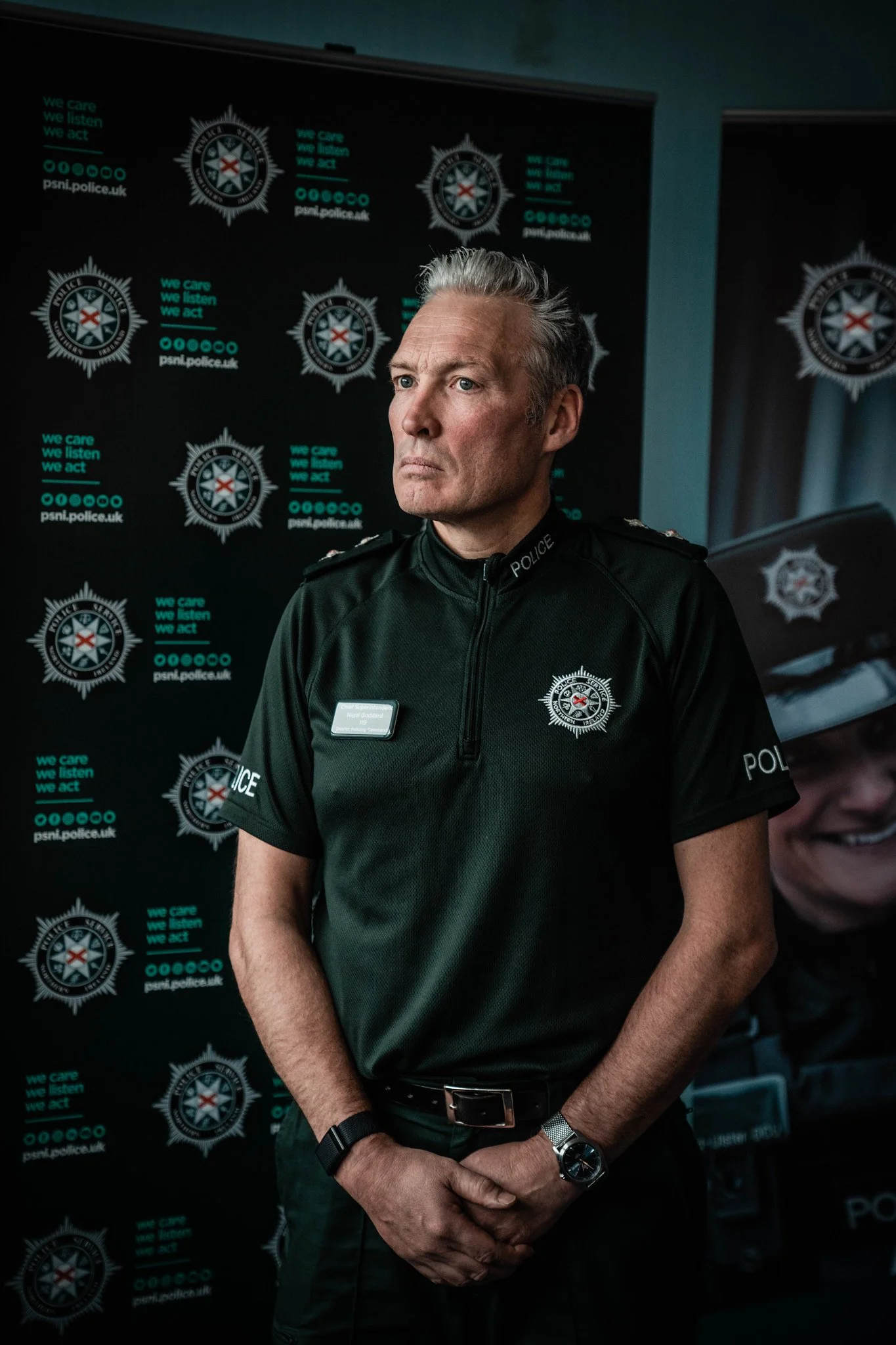 A male police officer with gray hair wearing a black police uniform and badge, standing in front of a backdrop with police insignia and the slogan "we care we listen we act."