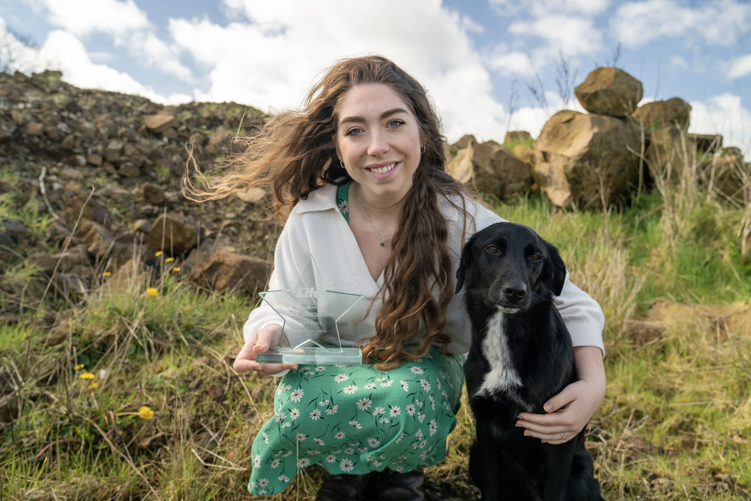 A woman with long brown hair smiling and holding a glass star-shaped award, kneeling beside a black dog with a white patch on its chest, outdoors on a grassy area with rocks and cloudy sky in the background.