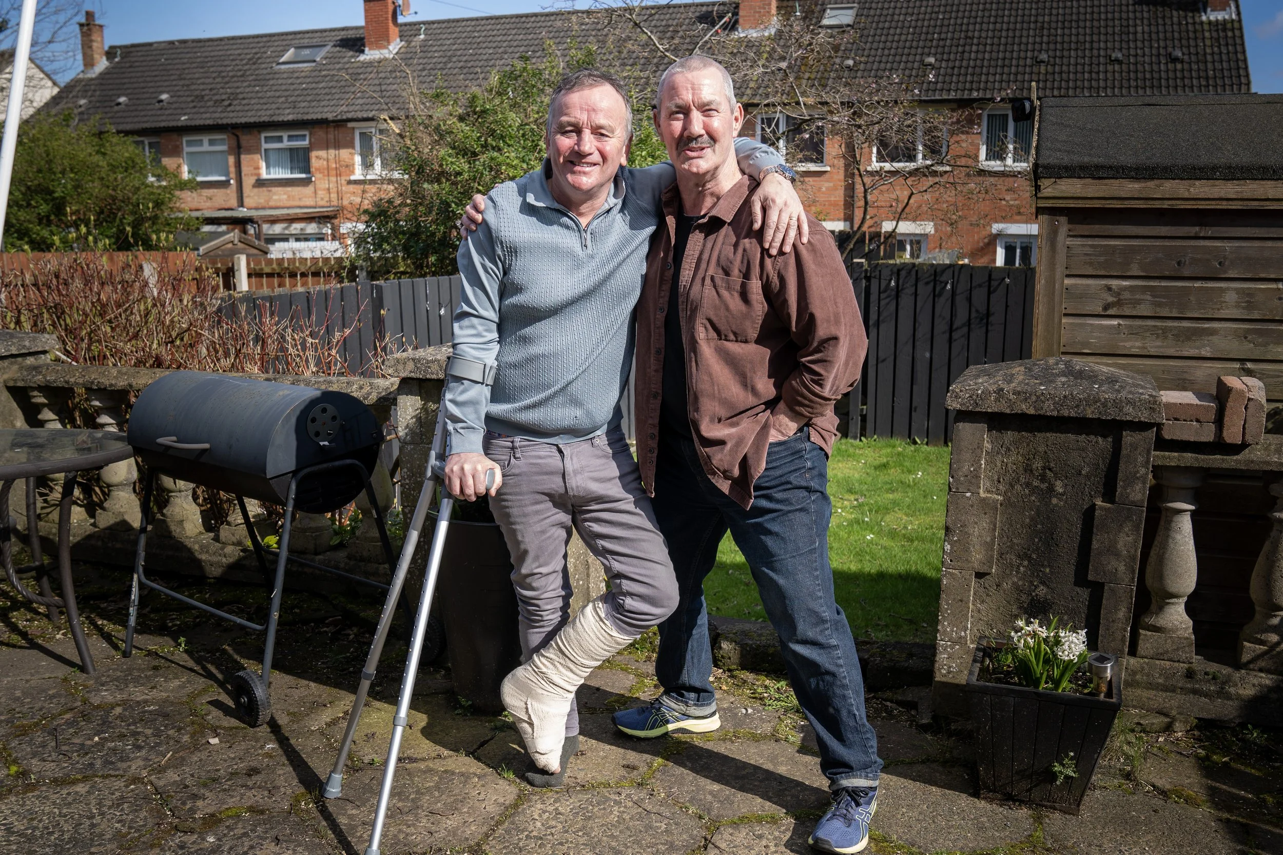 Two smiling men standing outdoors in a backyard, one in a cast on his leg with crutches, and the other with his arm around his friend. There is a barbecue grill beside them and houses in the background.