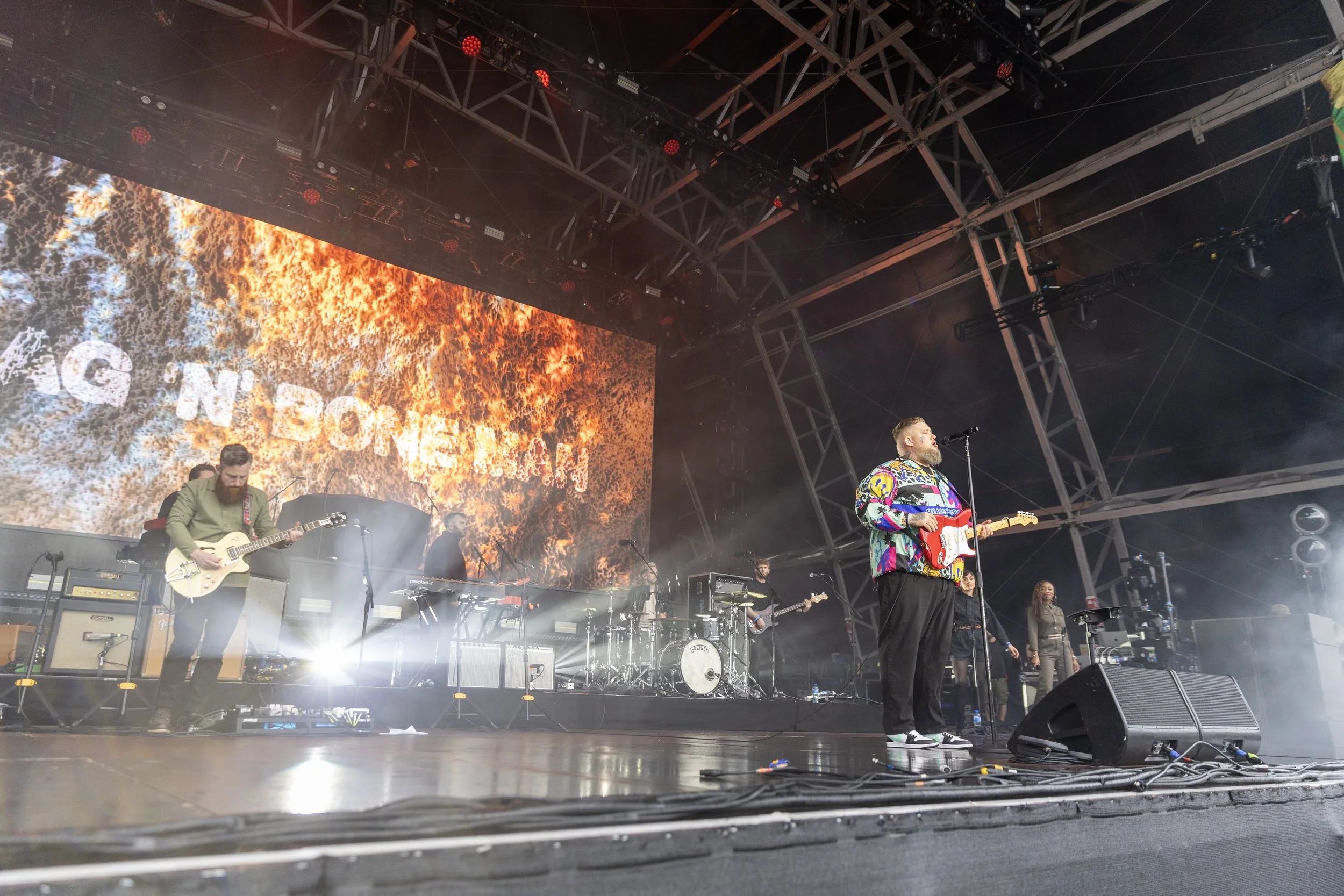 A band performing on stage with a large screen in the background displaying fiery graphics and partially visible text.