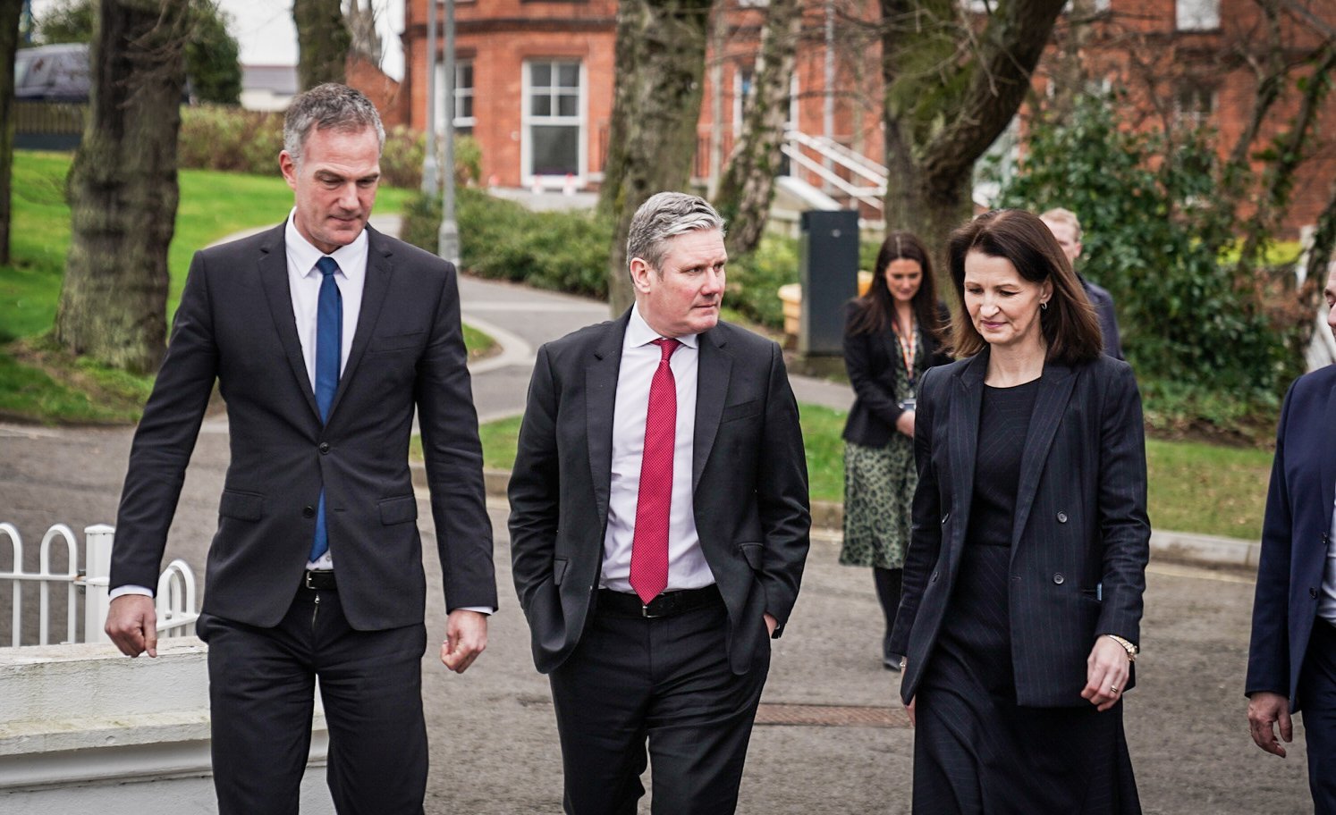Three professionally dressed individuals, two men and a woman, walking outdoors on a paved path, engaged in conversation. The background features trees, grass, and brick buildings.