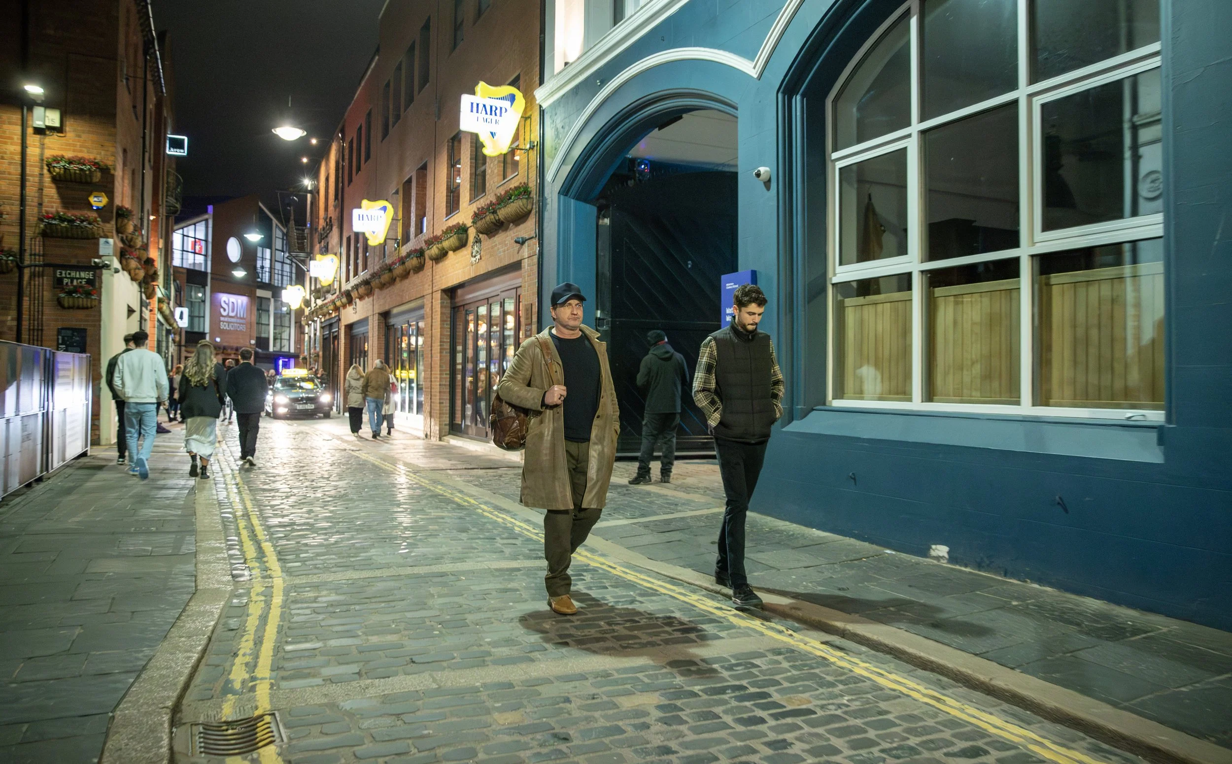 People walking along a city cobblestone street at night, decorated with hanging flower baskets and illuminated signs, including a Harp Lager sign.