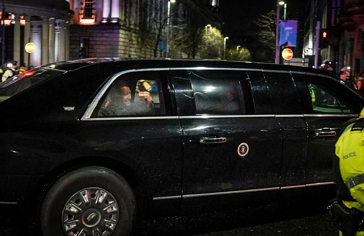 A black government limousine with a presidential seal on the door, parked on a city street at night, with police officers and others in the background.