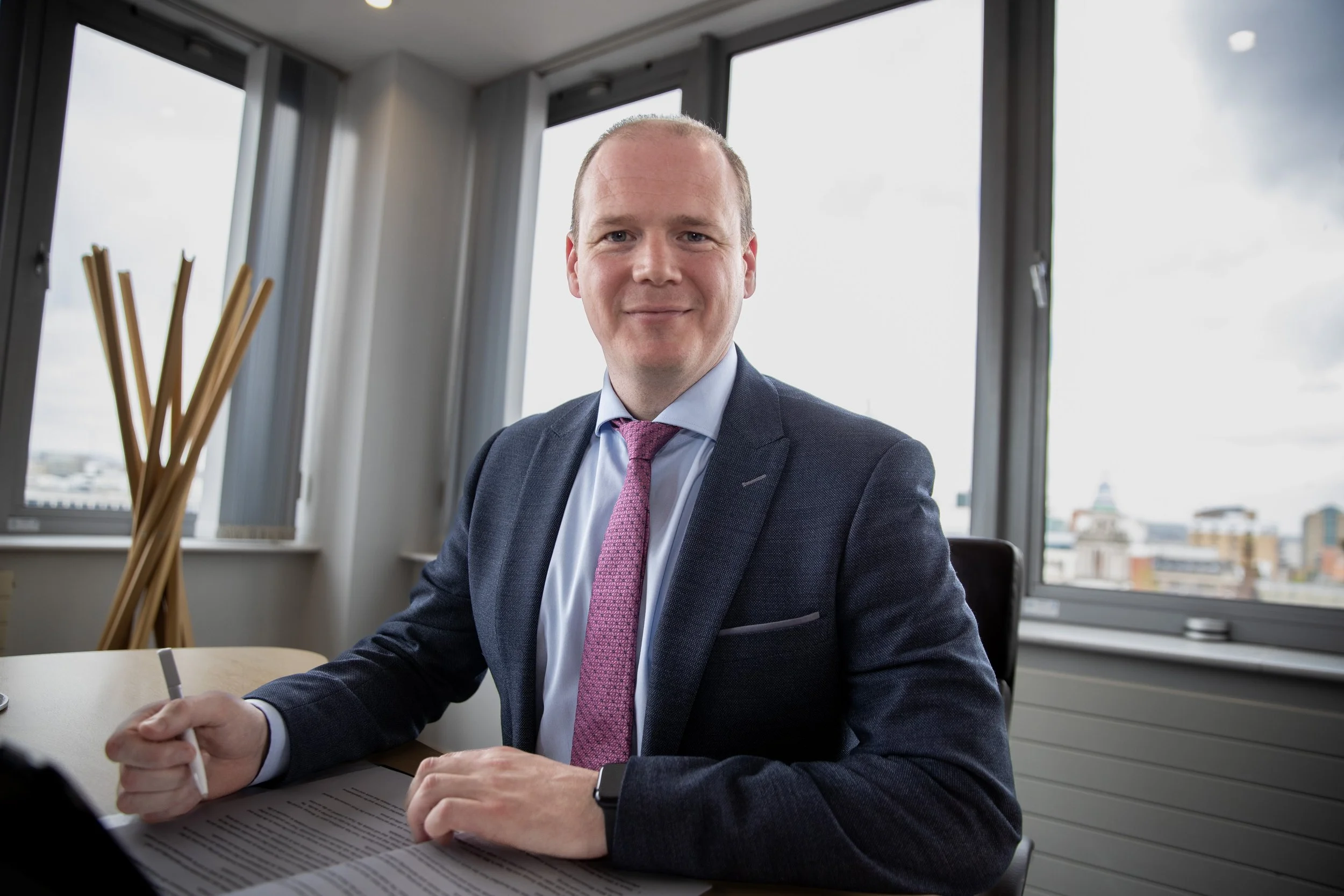 A man in a navy blue suit with a pink tie sitting at a desk in an office with large windows overlooking a cityscape, holding a pen and looking at the camera with a slight smile.