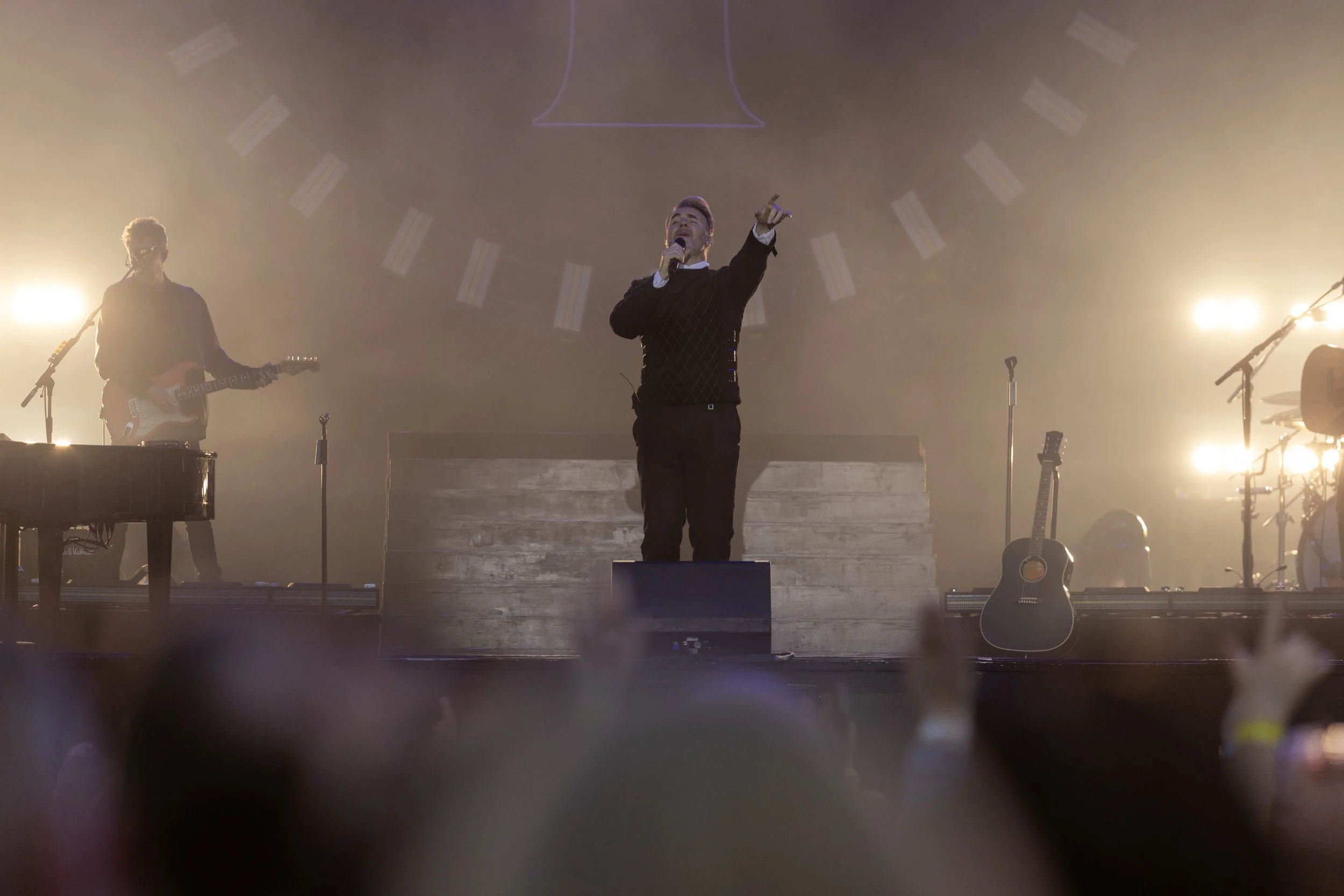 Performer singing on stage with a guitarist in the background and musical instruments, bright stage lights, and audience in the foreground.