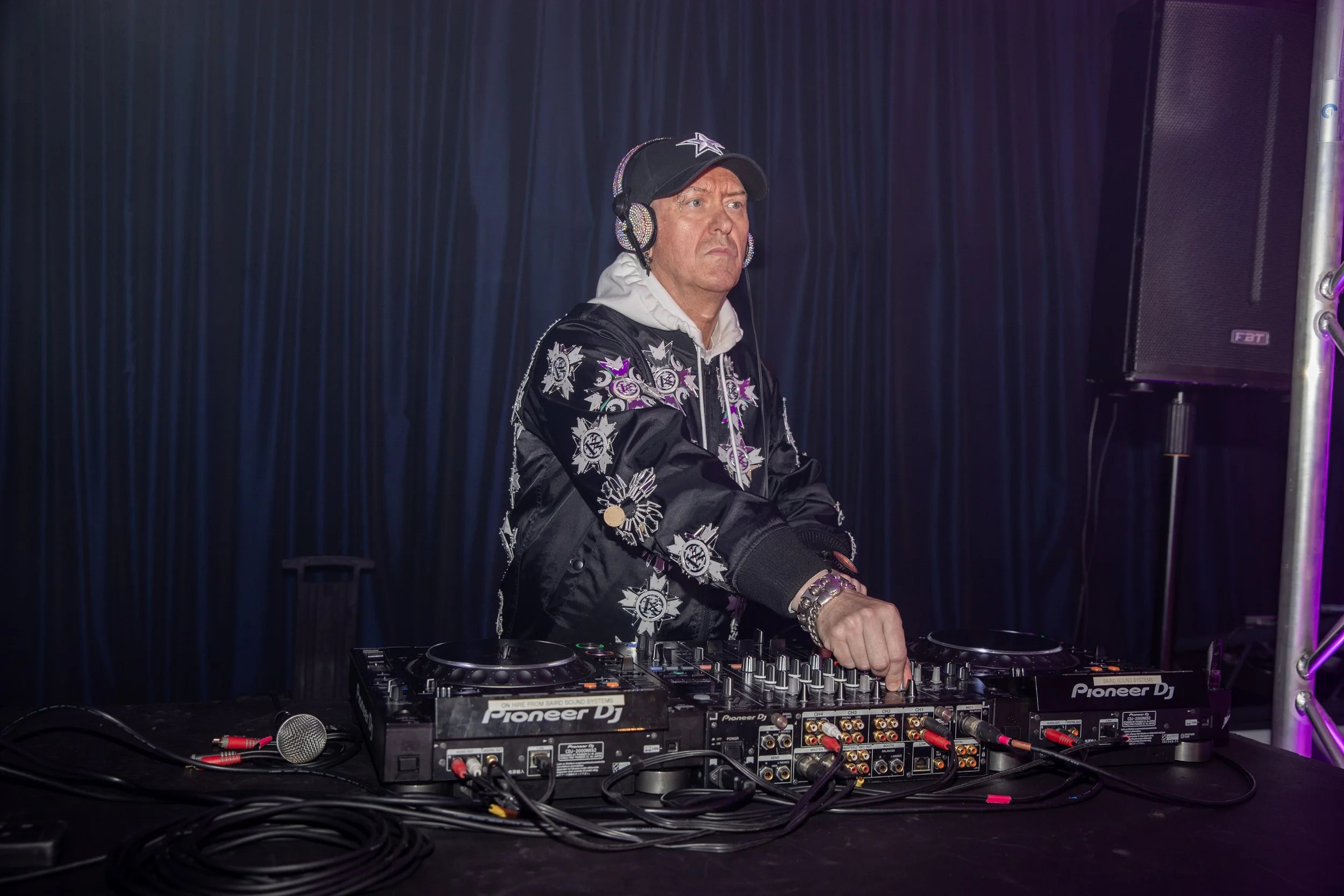 A DJ wearing a black cap, a hoodie, and a black jacket with purple and white graphics, standing at a Pioneer DJ setup with headphones on, in front of a dark blue curtain.