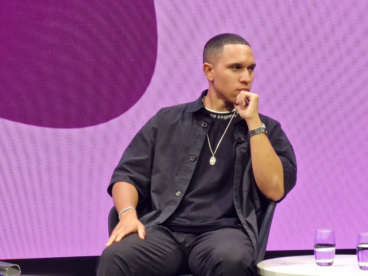 Young man sitting with his chin resting on his hand during an event, with a pink and purple background and two glasses of water on a table.