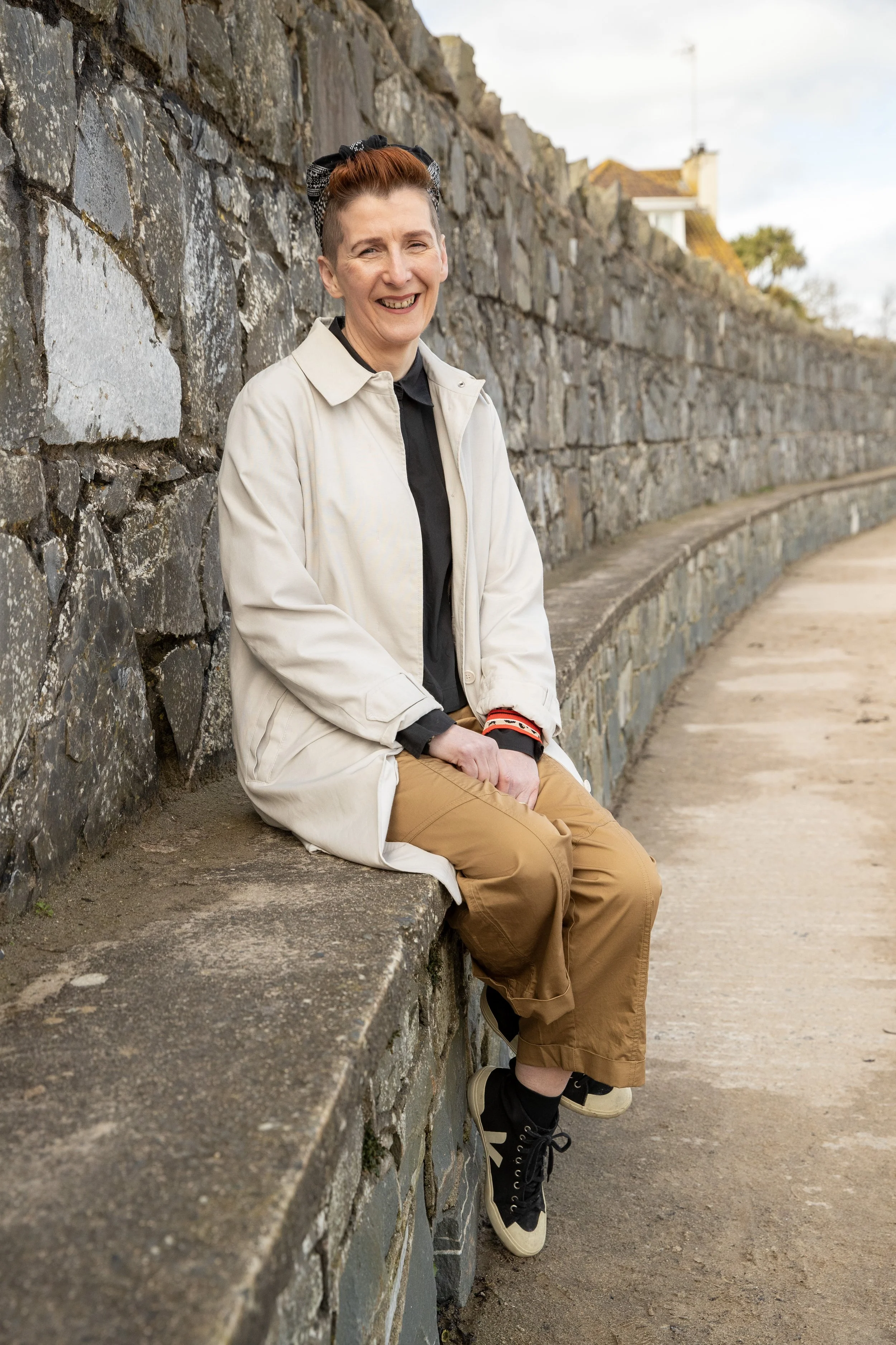 A woman with short, styled hair sitting on a stone wall outdoors, smiling, wearing a beige coat, black shirt, brown pants, and black sneakers.