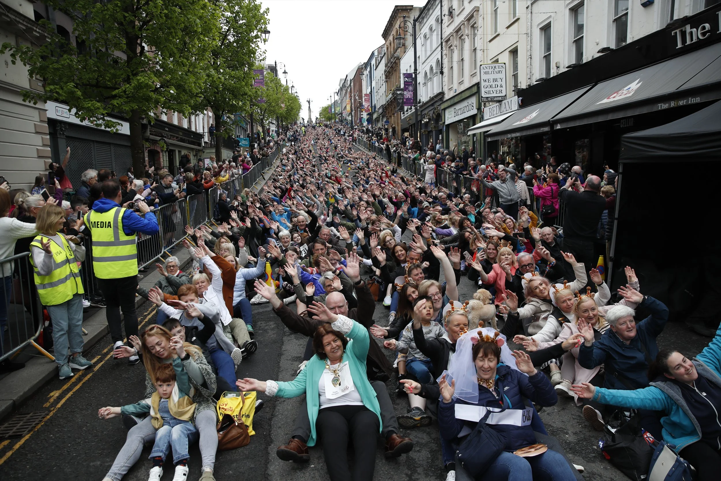 Large crowd of people sitting and kneeling on the street, raising their hands during a public event or parade, with spectators behind barriers on sidewalks and storefronts in the city.