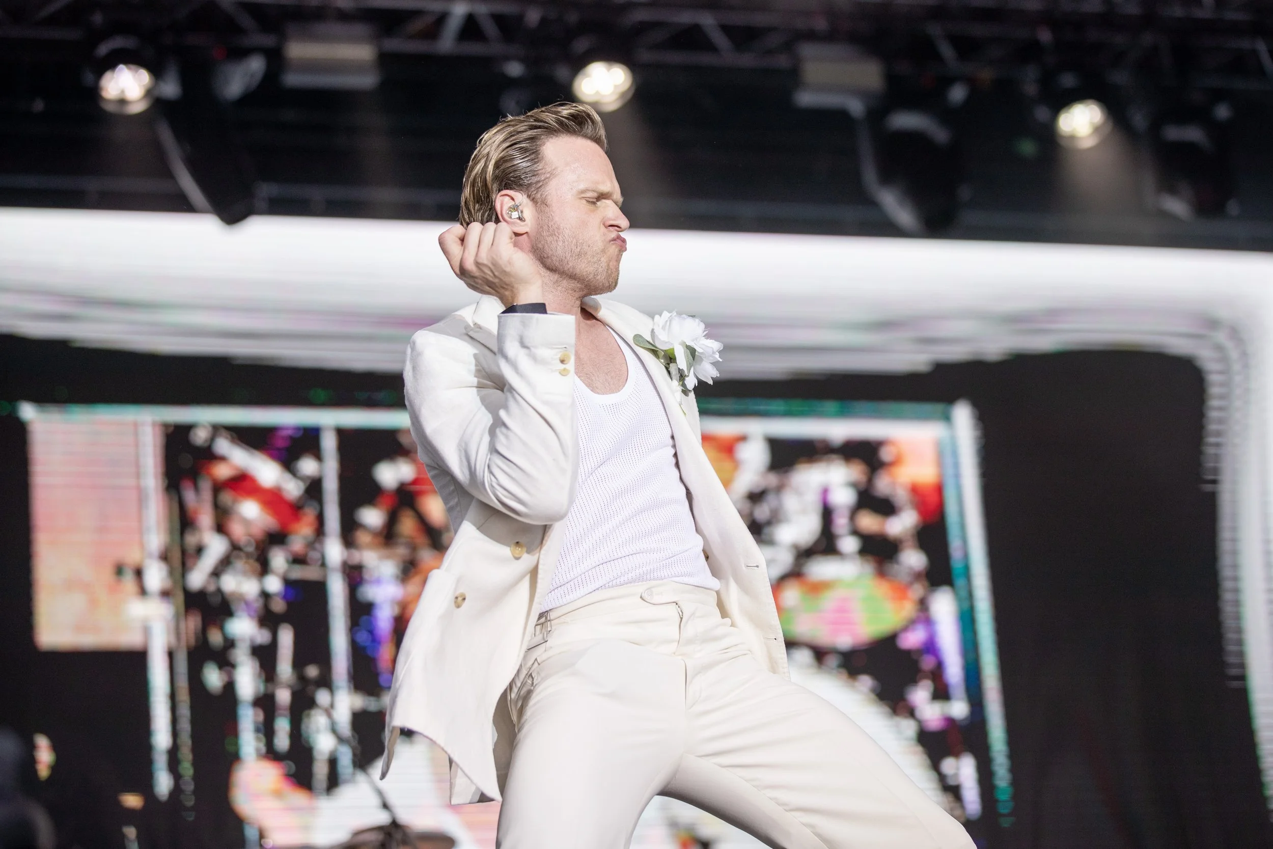 A male performer with styled blonde hair dancing on stage, dressed in a white suit with a boutonniere, making a pouty face with eyes closed, in front of a colorful digital background.