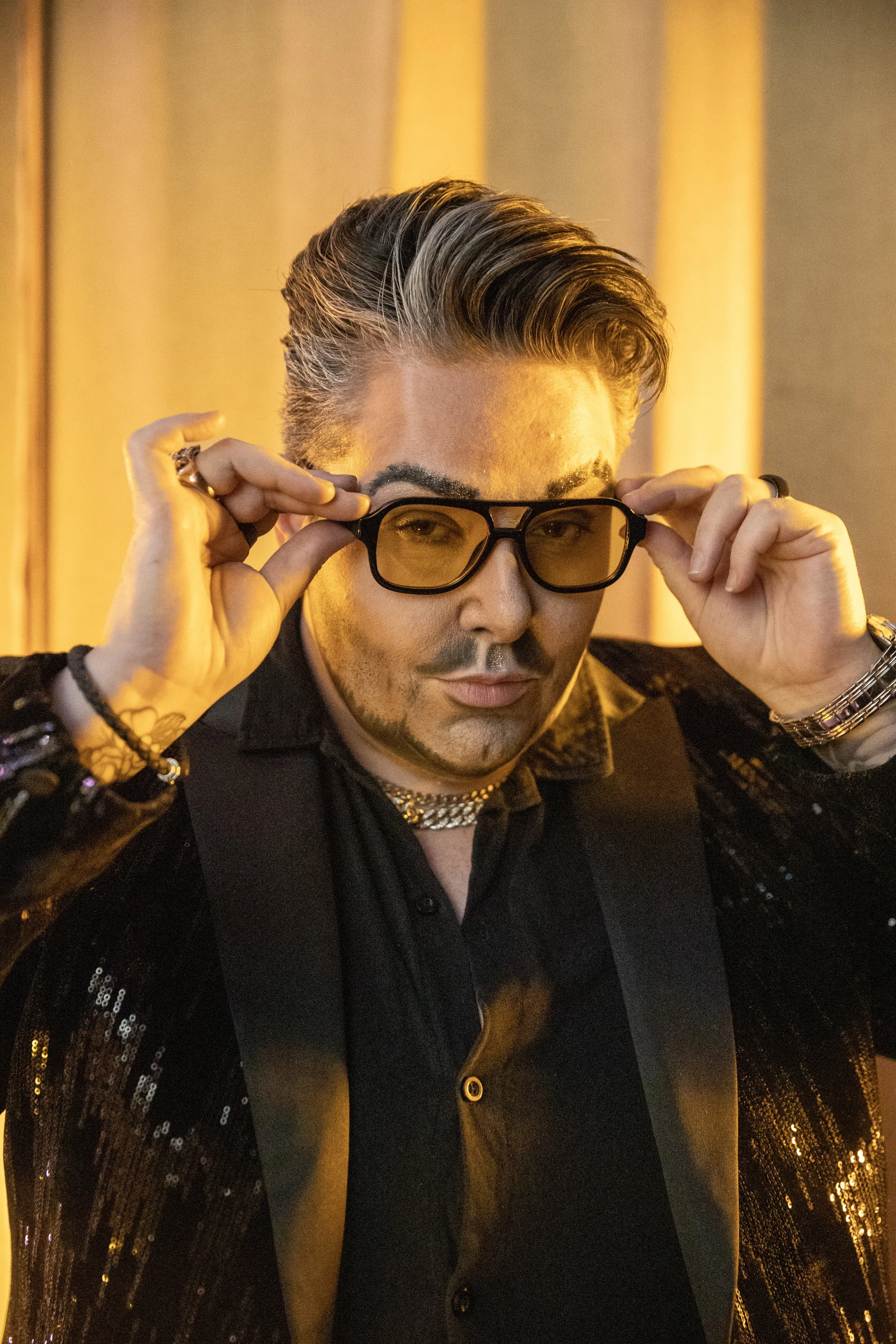 A young man with styled hair, wearing black-rimmed glasses, a black shirt, and a shiny black blazer, holding his glasses and looking directly at the camera.