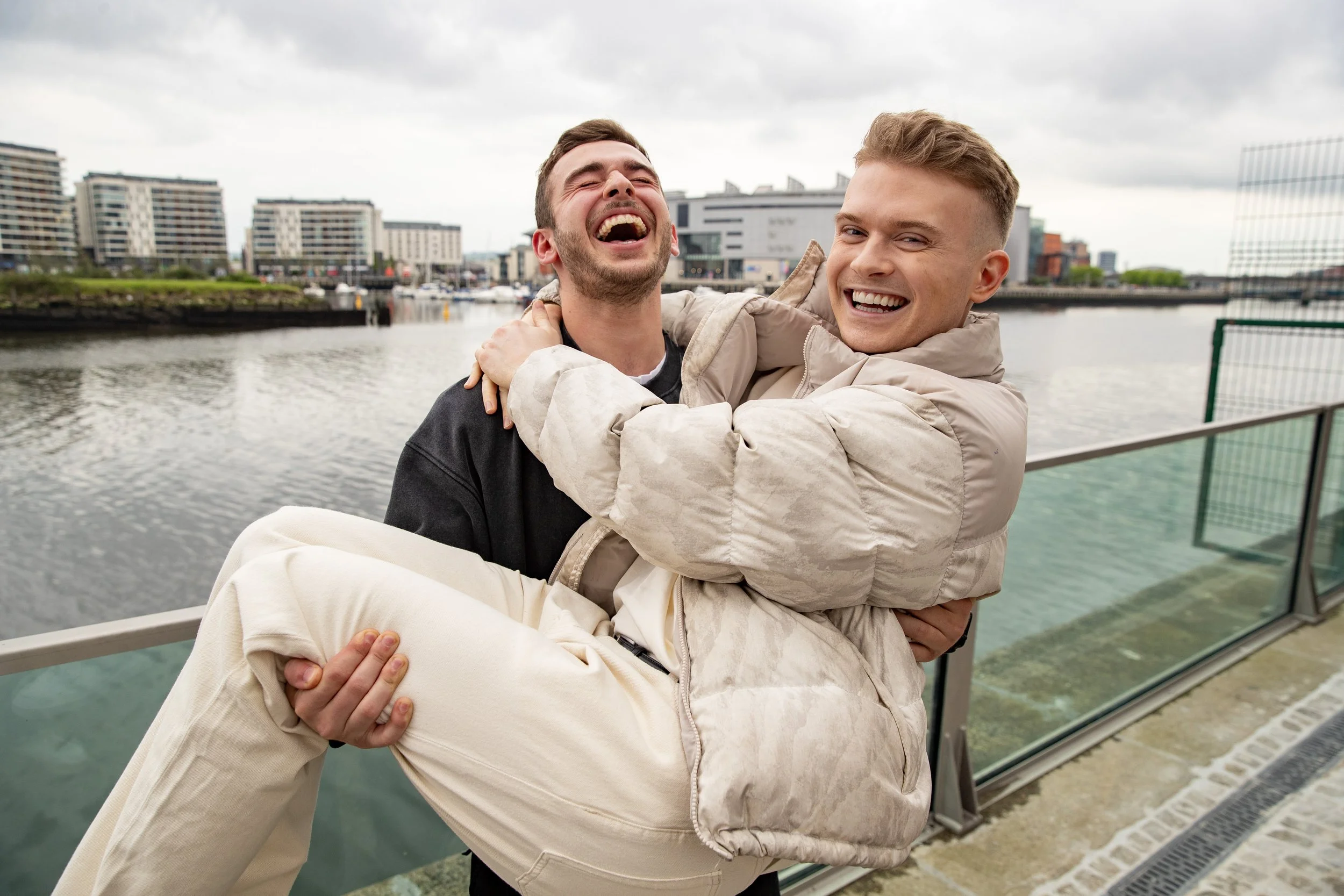 Two young men smiling and laughing, one holding the other in his arms, standing by a body of water in an urban area with modern buildings in the background.