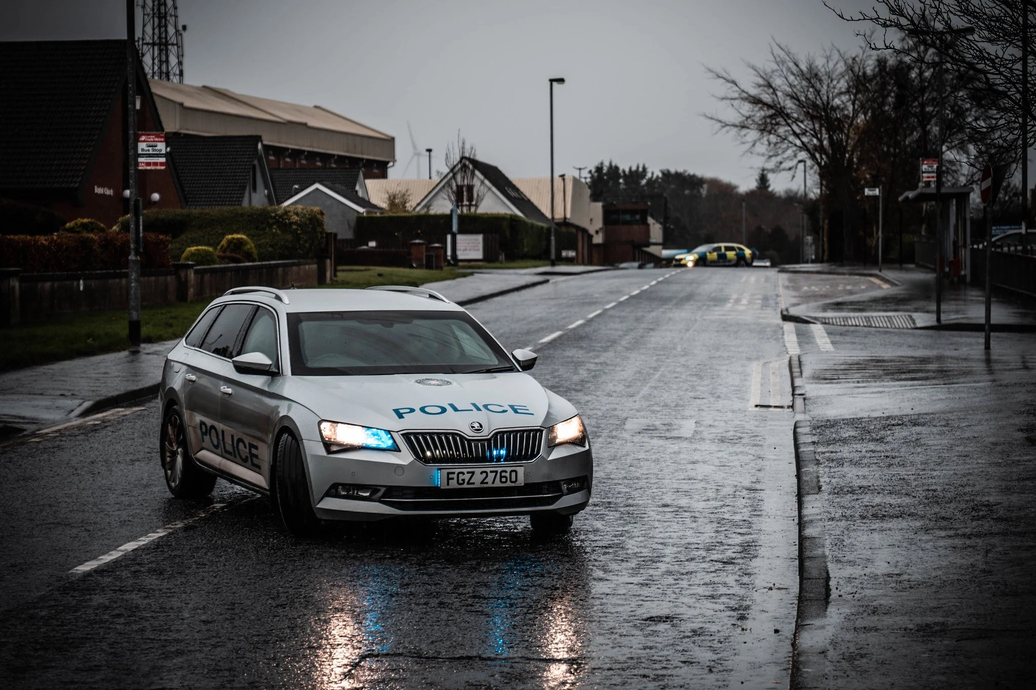 Police car with flashing lights parked on wet road in a residential area during overcast weather, with other police vehicles in the background.