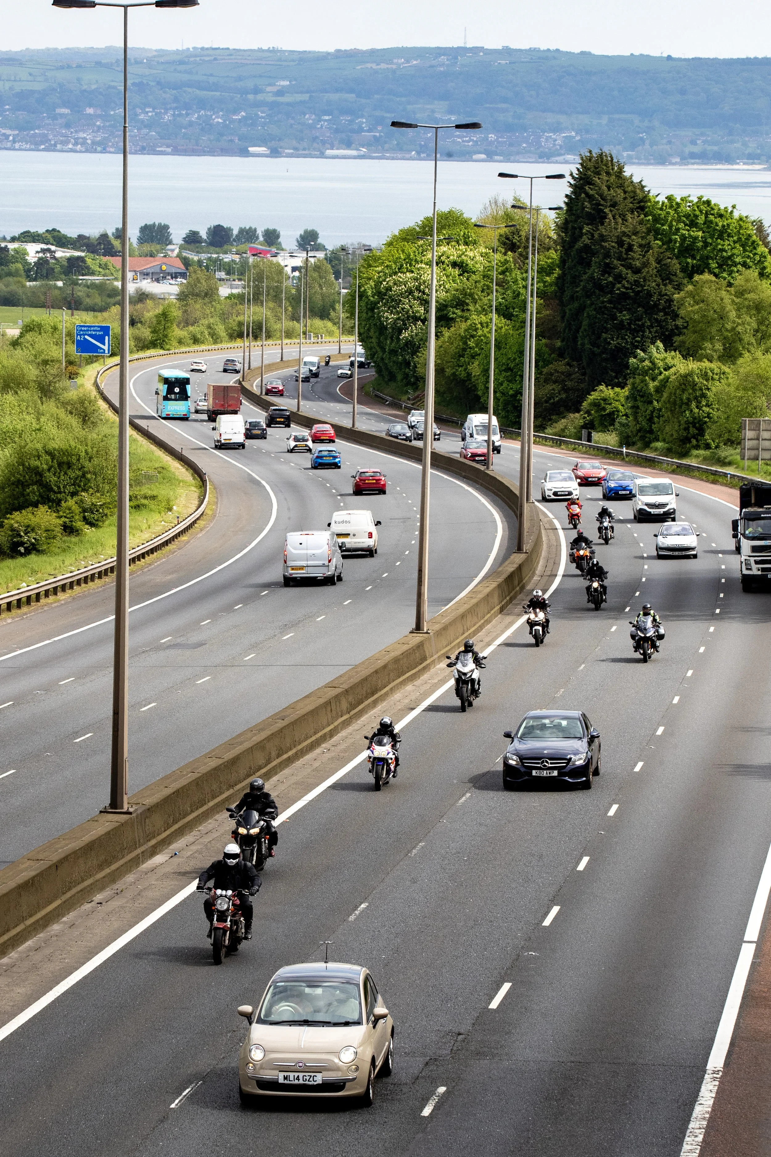 Multiple cars and motorcycles driving on a multi-lane highway with green trees on the side and water in the background.