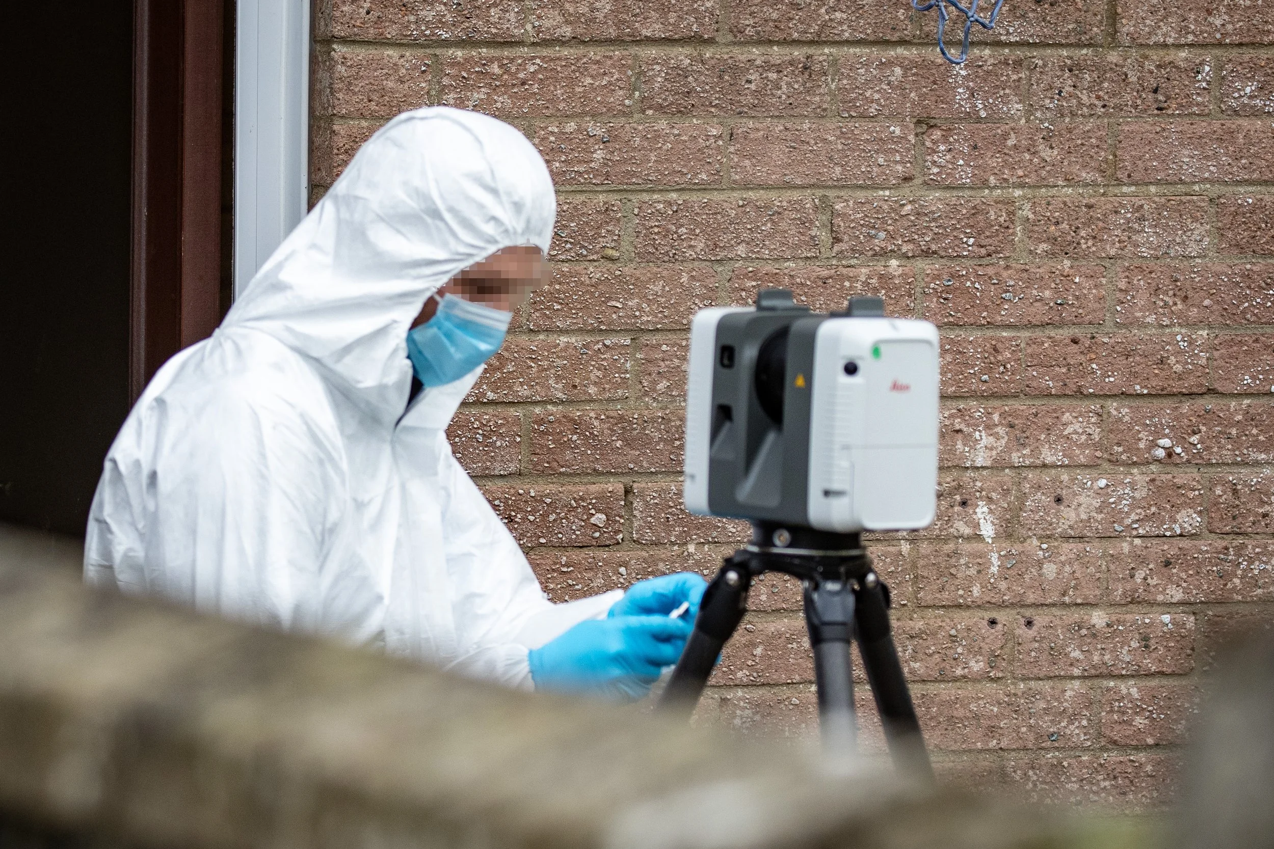 Person wearing protective suit, mask, and gloves using specialized equipment outdoors next to a brick wall.