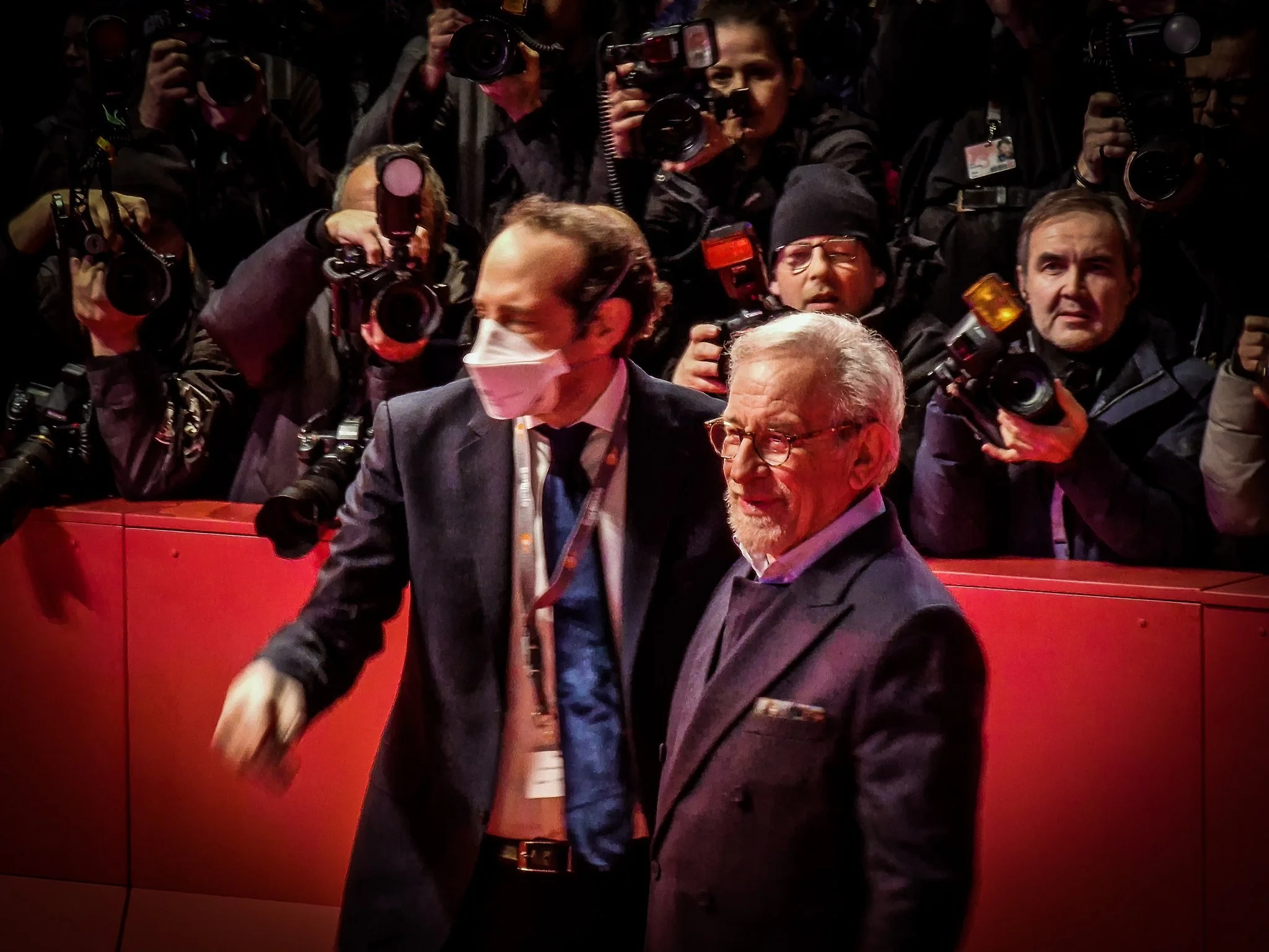 Two men walking past a crowd of photographers, one wearing a face mask and glasses, the other with gray hair and a beard, both dressed in suits, with the crowd taking pictures of them.