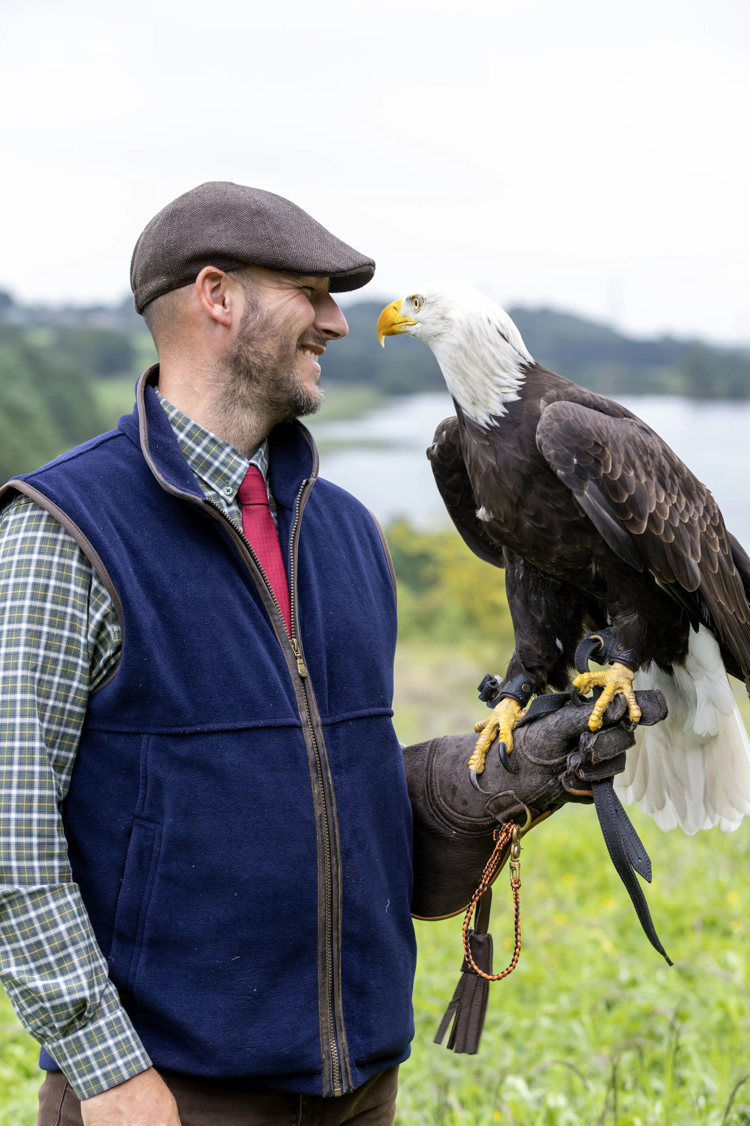 A man in a brown cap, plaid shirt, red tie, and navy vest smiling at a bald eagle perched on his gloved hand outdoors with a lake and green landscape in the background.