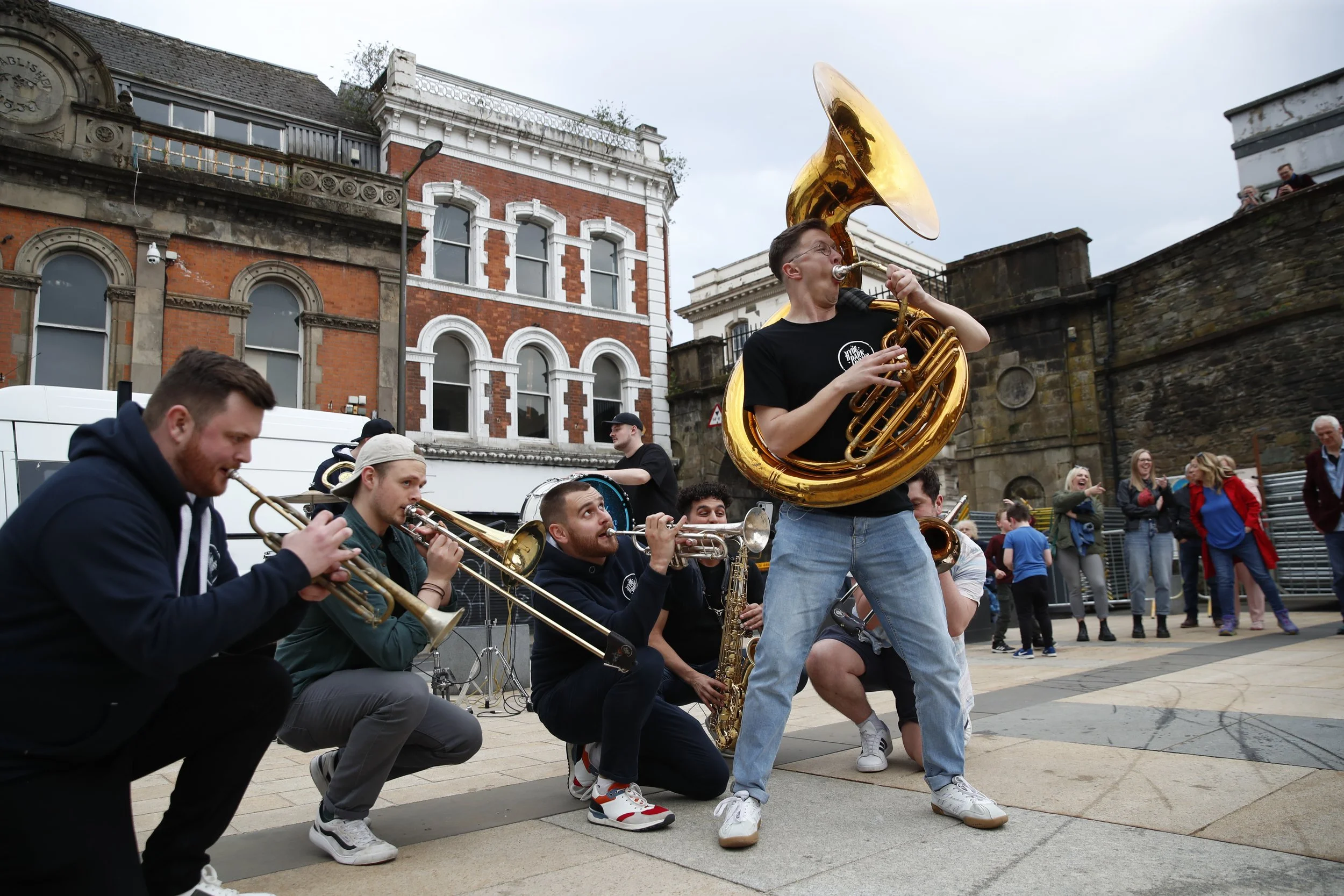 A street band performing outdoors with a man playing a sousaphone in the front, and other musicians playing trumpets, trombones, and saxophones behind him, while onlookers watch and smile.