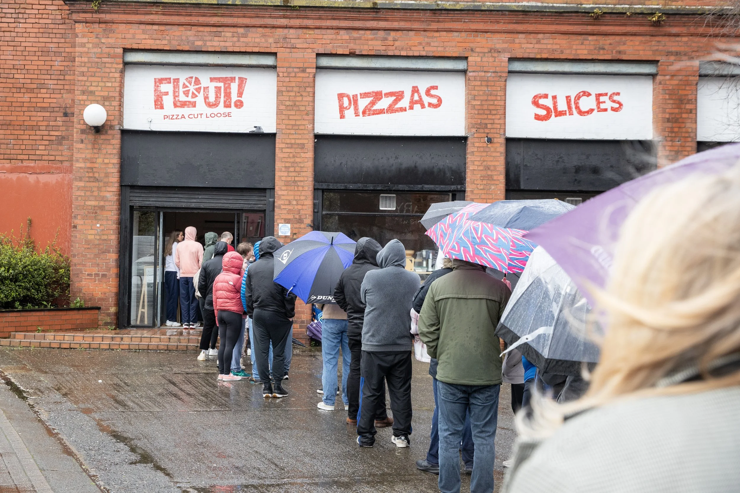 People standing in line outside a pizzeria in rainy weather, holding umbrellas. The pizza shop has signs that say 'FLOUT! PIZZAS SLICES' and is in a brick building.