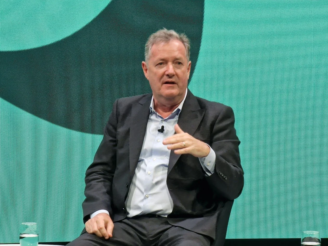 A middle-aged man wearing a dark suit and light blue shirt is seated, speaking during a panel discussion or interview. There are two glasses of water on the table in front of him. The background features a large screen with a turquoise and black grap