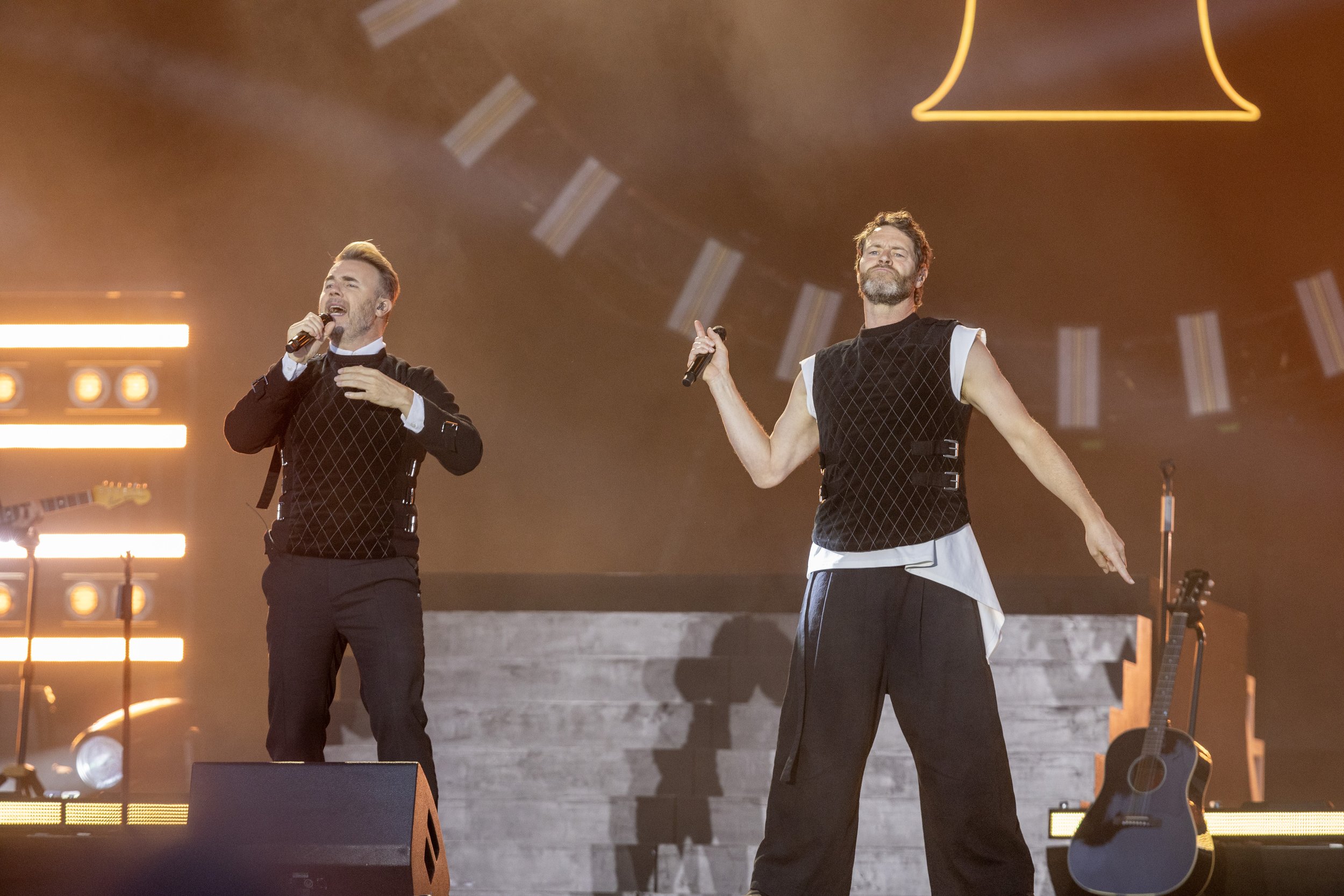 Two men singing on stage during a concert, with one holding a microphone and the other gesturing with his right hand. There are musical instruments and stage lights in the background.