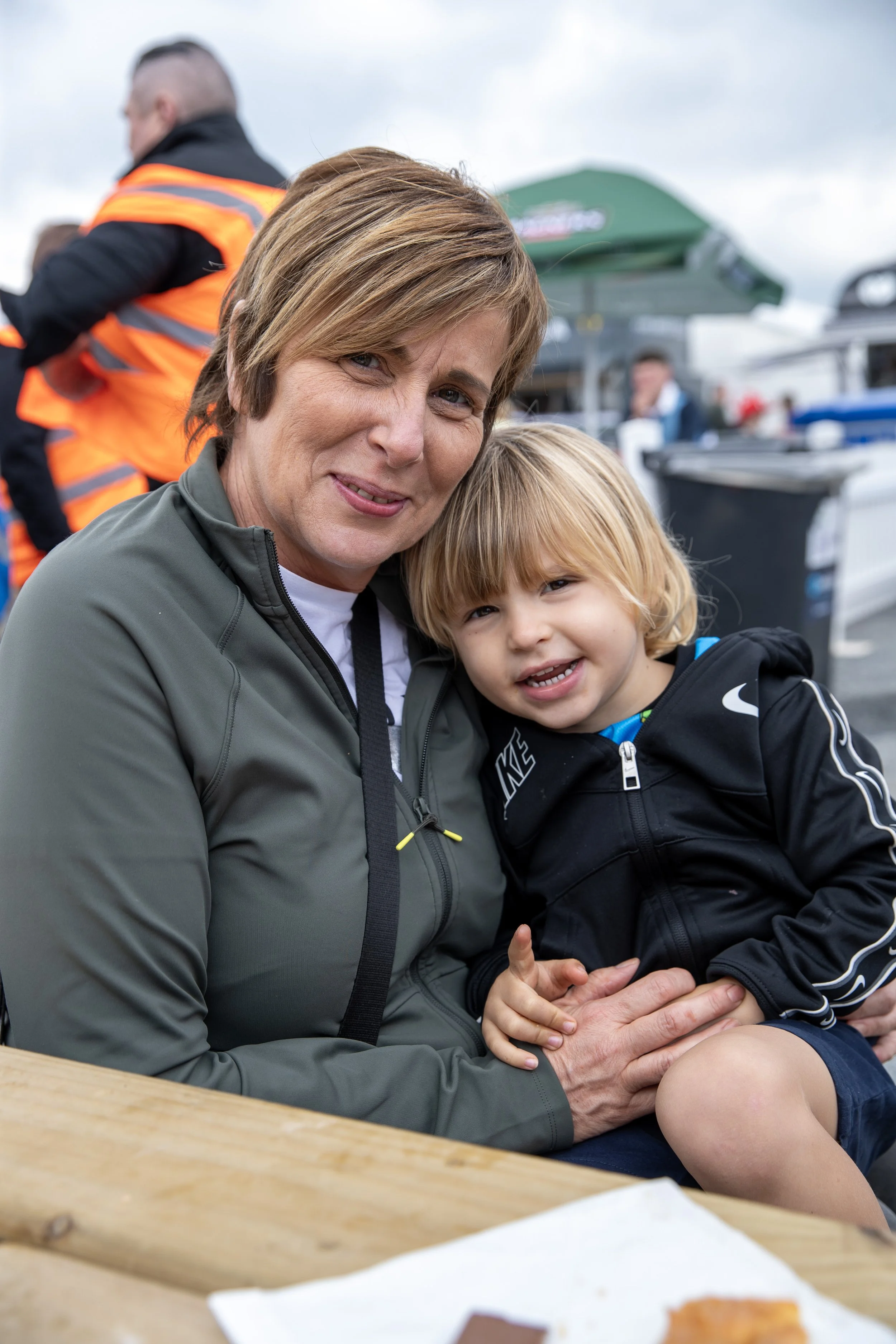 A woman and a young boy are sitting together outdoors, smiling at the camera. The woman has short brown hair and is wearing a gray jacket, while the boy has blond hair and is wearing a black Nike jacket. There are people and boats in the background.