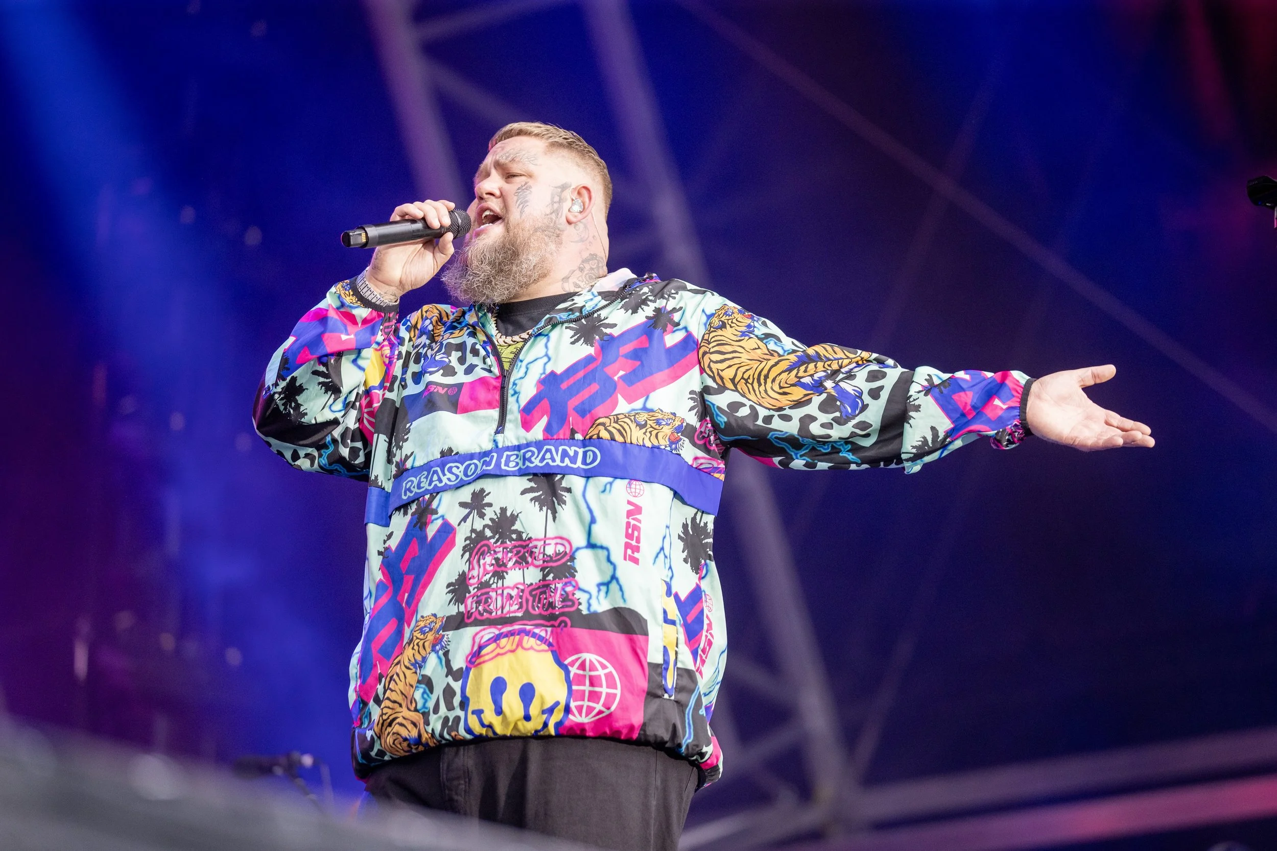 Male performer with a beard singing into a microphone on stage, wearing a colorful, patterned jacket featuring tigers, skulls, and palm trees.