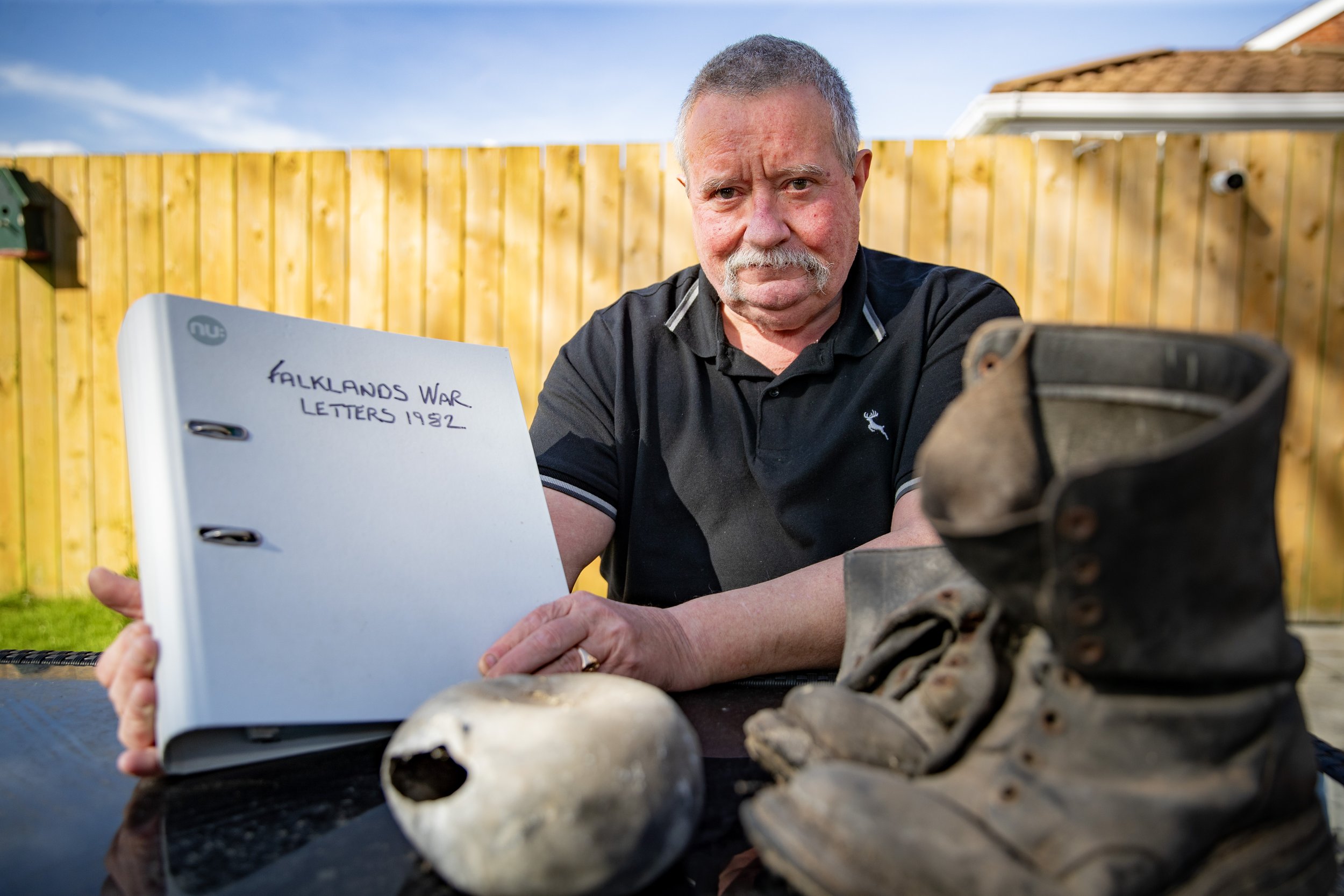 An older man with a mustache sitting outdoors at a table, holding a binder that reads 'Falklands War Letters 1982,' with a skull and worn boots on the table in front of him, and a wooden fence and blue sky in the background.