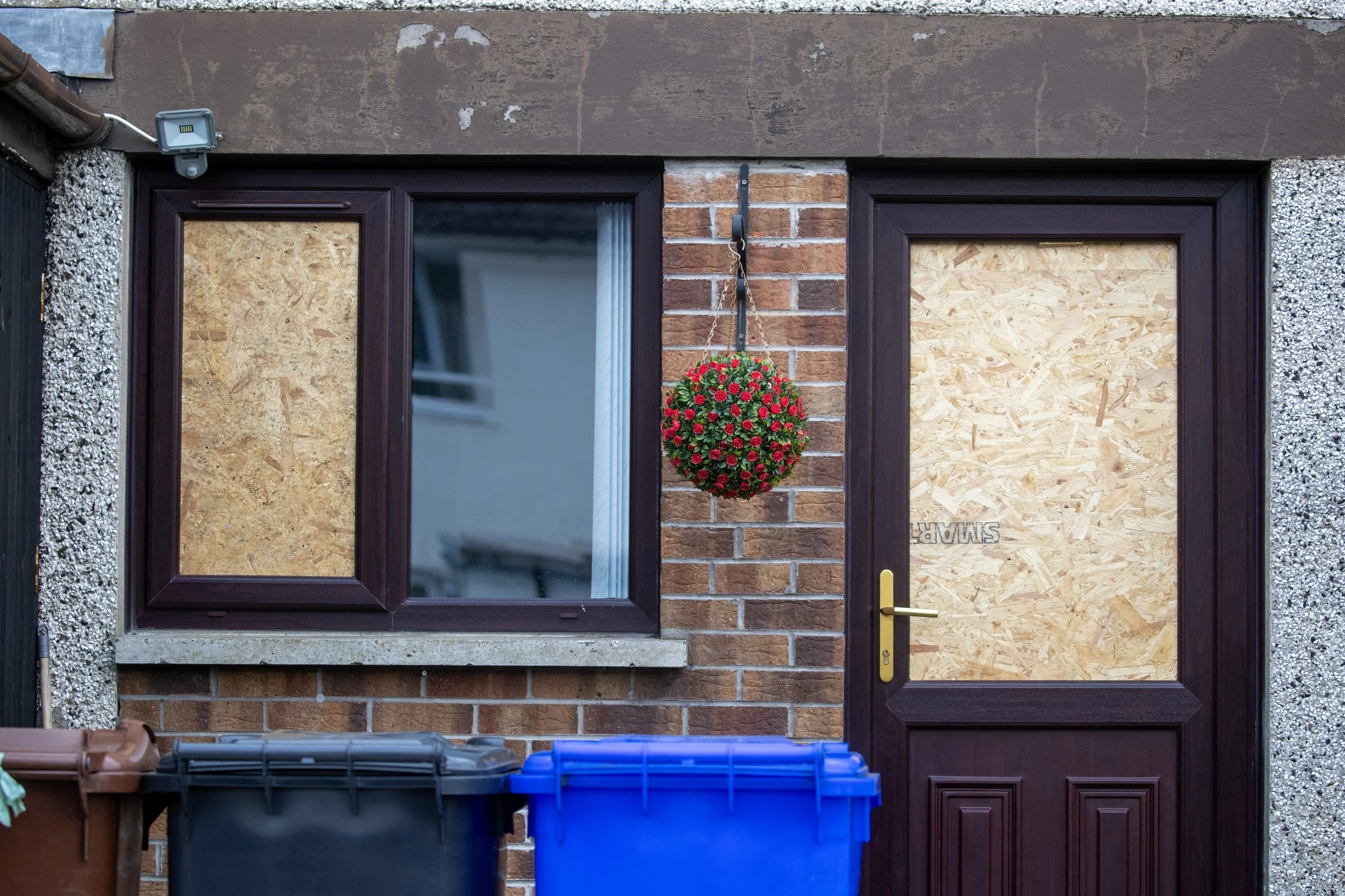 Front of a house with boarded-up windows and door, a hanging flower basket in the center, and three trash bins in front.