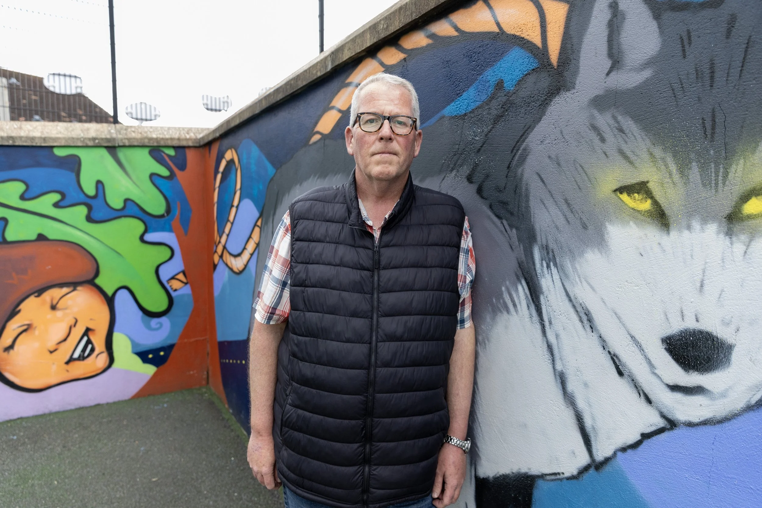 A man with gray hair and glasses standing in front of a colorful mural of a wolf with yellow eyes and a child's face sleeping on a tree branch.